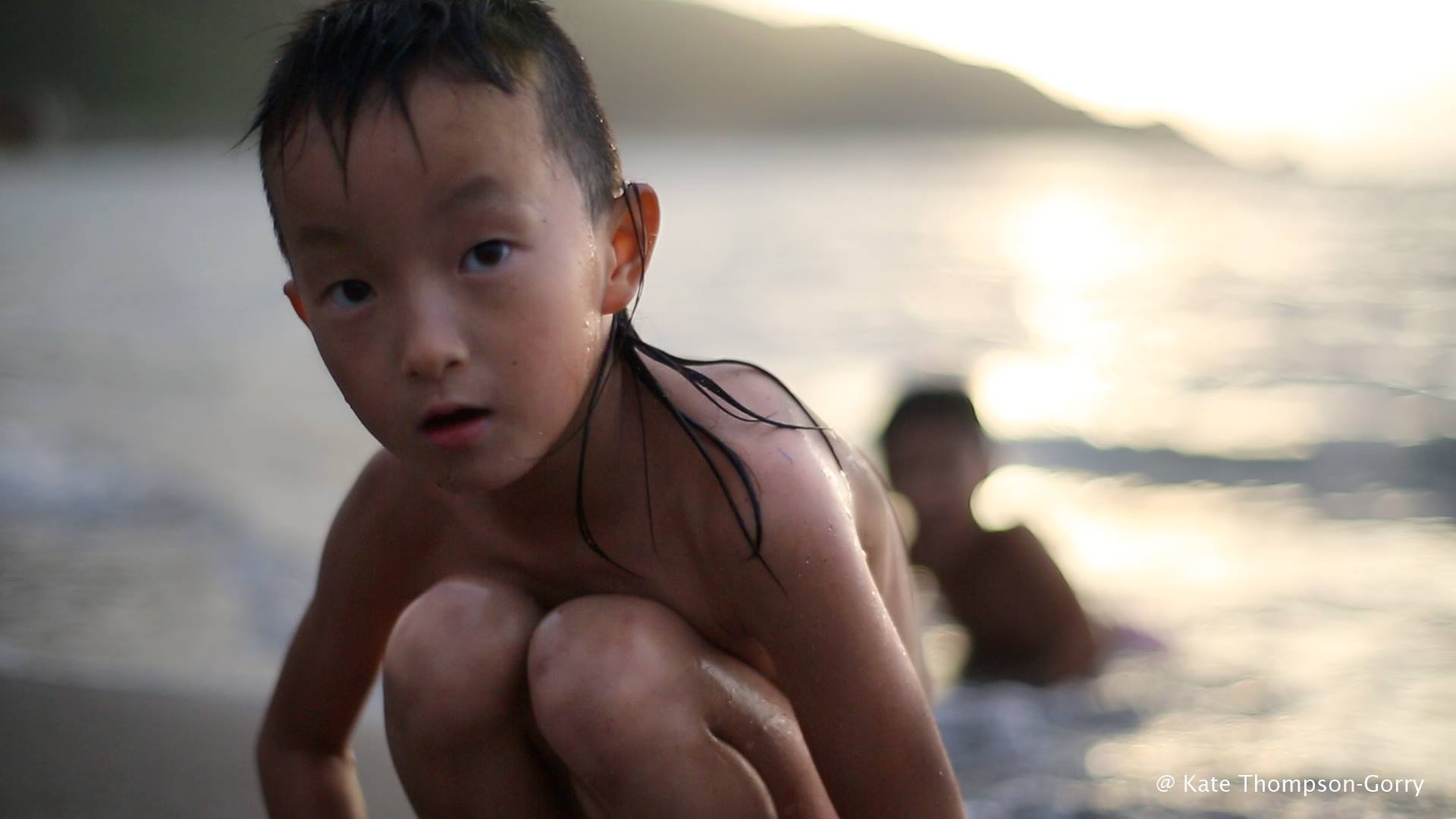 A young child with wet hair crouches on a beach, looking toward the camera. The sunset casts a warm glow on the water and mountains in the blurred background. Another child is swimming in the shallow water behind them. Photo credit: Kate Thompson-Gorry.