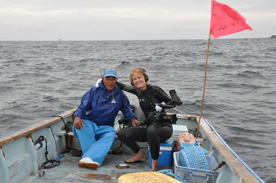 A man in a blue outfit and a woman in a wetsuit are sitting together on a small boat in the ocean. The woman is holding a camera. There is a pink flag at the back of the boat, and the sky is overcast. Various baskets and equipment are visible on the boat.