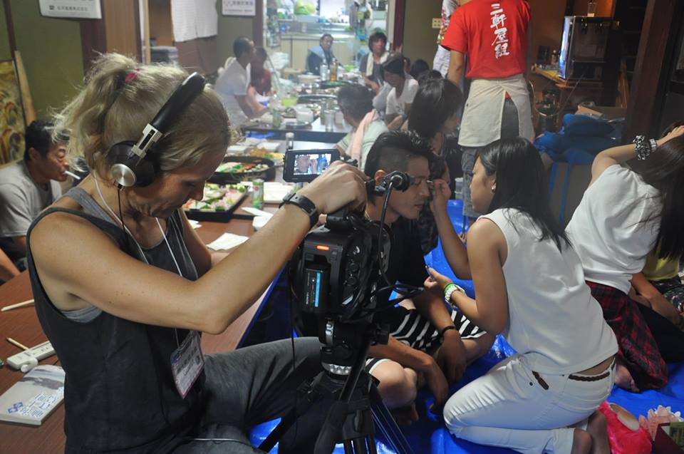 A person wearing headphones and operating a camera films a group of people in a room. One person in the foreground is getting makeup applied. The room is filled with other individuals engaged in various activities, and there is food spread out on tables.