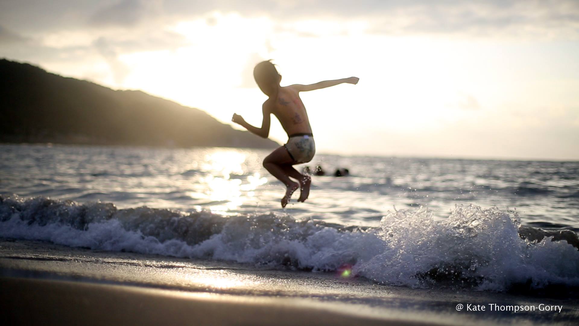A child leaps into the air over a small wave at the beach during sunset. The silhouette of a mountainous coastline is visible in the background, and the ocean sparkles in the sunlight. The child is wearing swim shorts and appears to be in mid-jump. © Kate Thompson-Gorry.