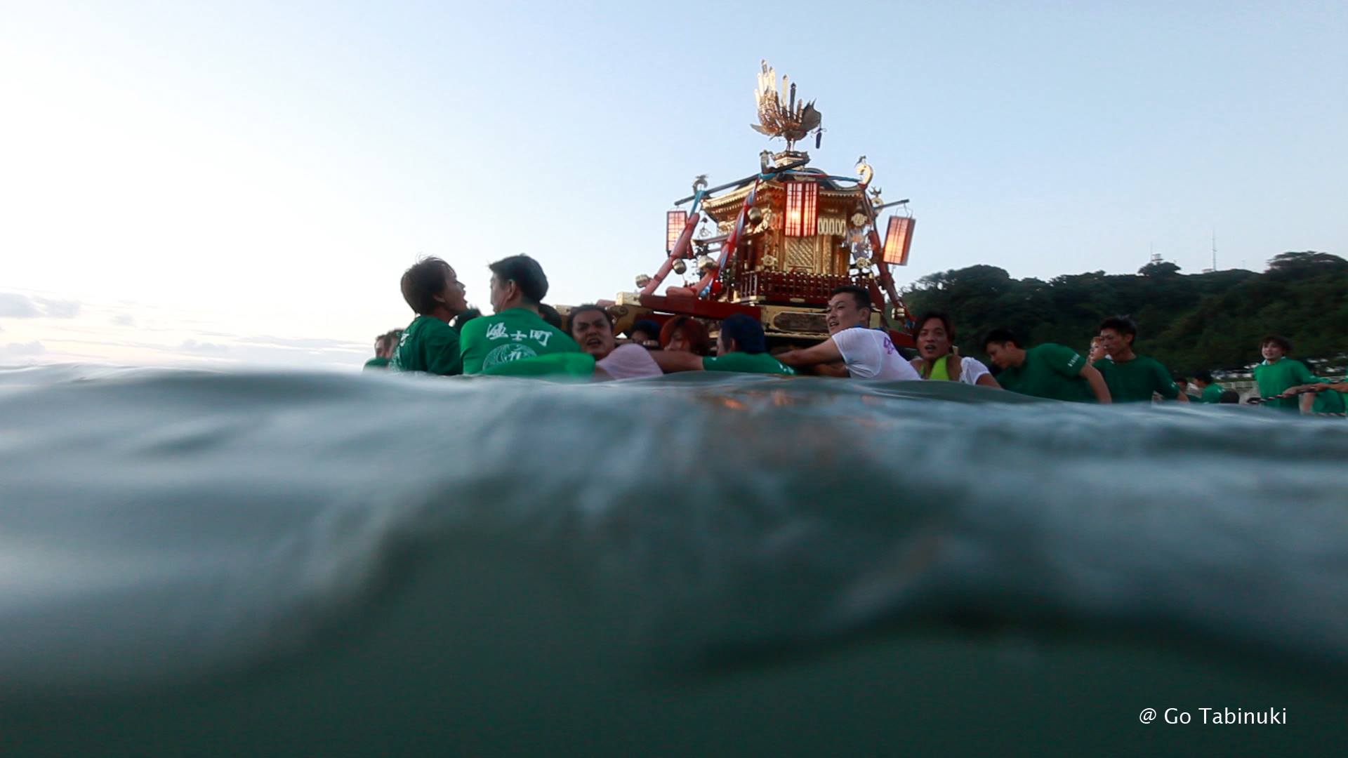 A group of people wearing green shirts carry a decorated palanquin through the water during a festival. The sky is clear and light blue, and a wooded hill is visible in the background. The water is partially obscuring the lower part of the group. © Go Tabinuki.