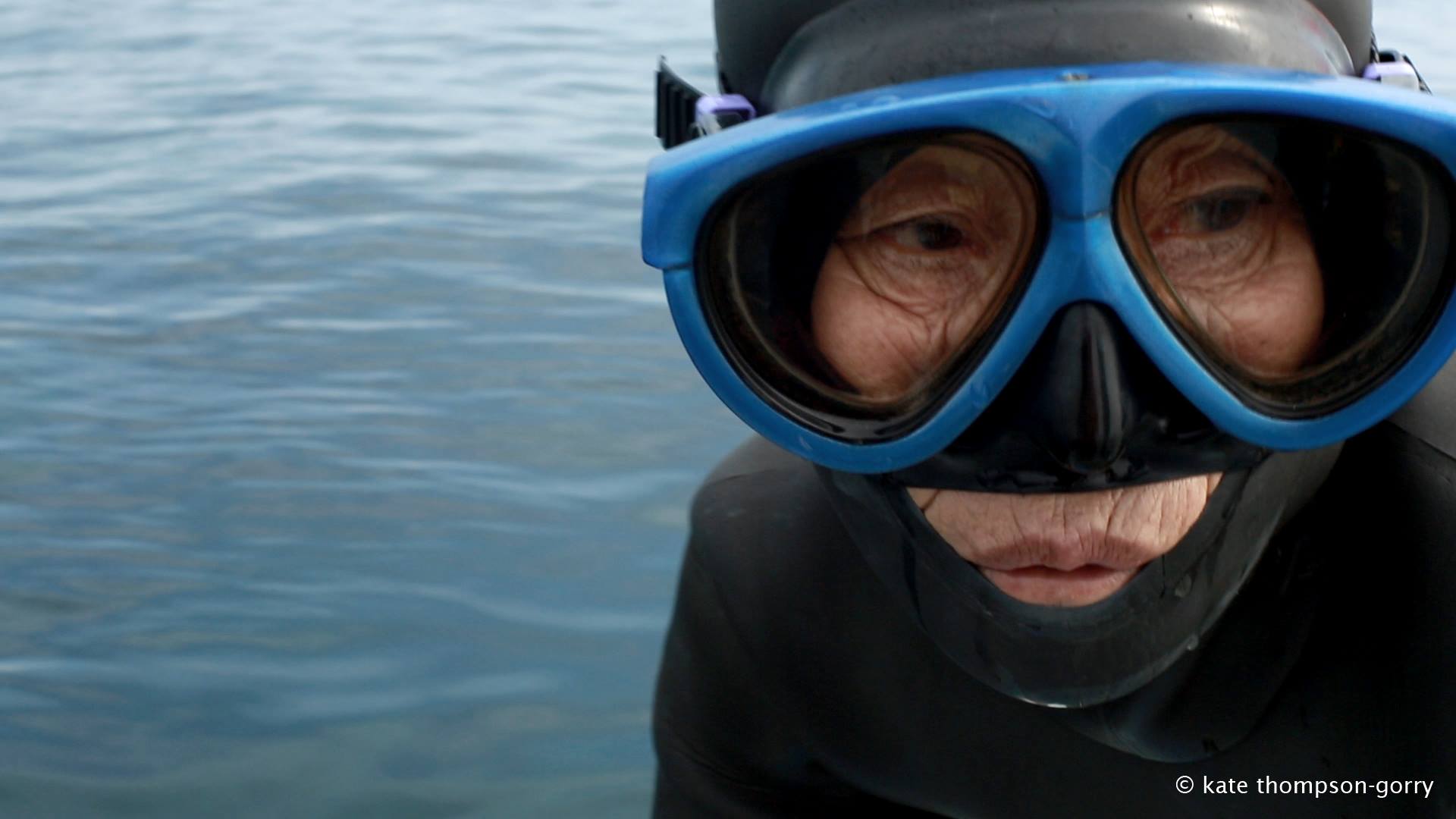 A person wearing a diving mask and wetsuit is partially submerged in water. The diving mask has blue trim, and the person is looking downward. The water is calm and reflects a light blue color. (© Kate Thompson-Gorry).