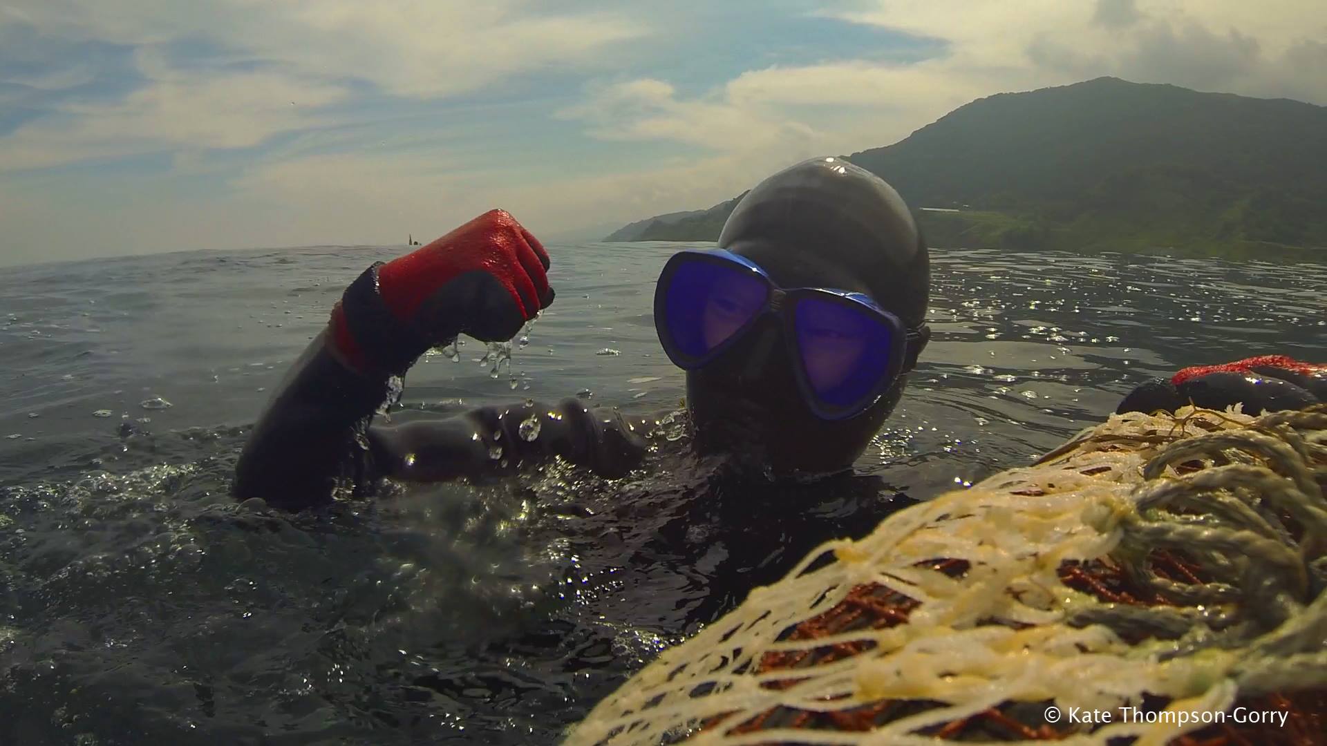A person wearing a black wetsuit, red gloves, and blue diving goggles is swimming in the ocean near a net. The individual is slightly submerged, with one hand raised, and water droplets splashing around. A coastal landscape is visible in the background. © Kate Thompson-Gorry.