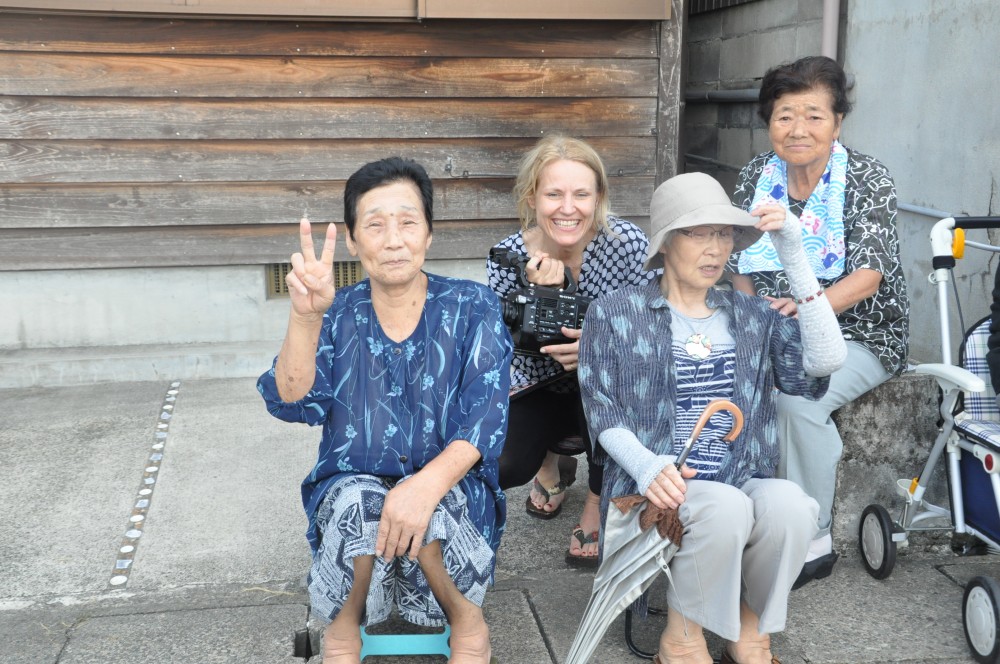 A group of four elderly women and a younger woman smile for the camera. One woman is kneeling with a peace sign, another holds a white cane and an umbrella while seated, a woman in a wheelchair is nearby, and the younger woman crouches in the middle holding a camera.