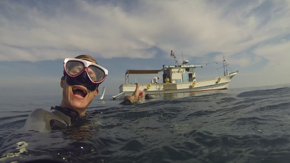 A person wearing snorkeling gear, including goggles and a breathing tube, smiles and points towards a boat in the background while floating in the open water. The boat is white with a red canopy and other equipment on board. The sky is partly cloudy.