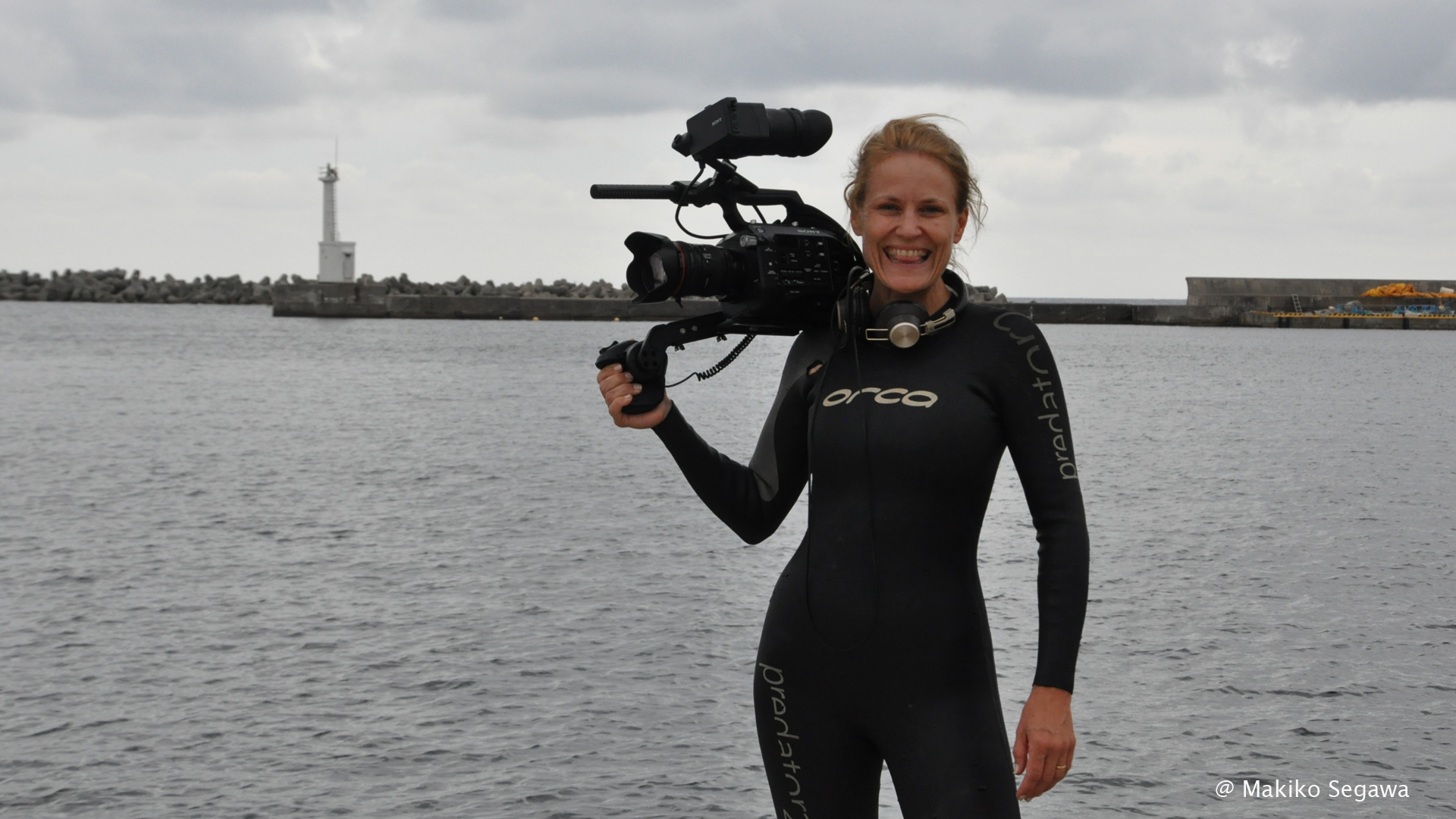 A smiling person in a black wetsuit holds a camera standing by a body of water with a lighthouse and breakwater visible in the background under a cloudy sky. ©Makiko Segawa is noted in the corner.