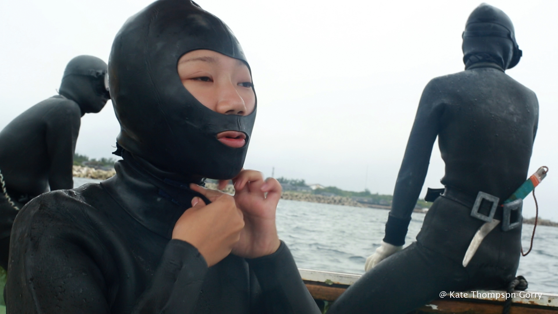 A person in a black wetsuit and diving hood adjusts the collar while sitting on a boat. Another person in similar gear sits facing the water, and a third appears in the background. The ocean and a distant shore are visible.
