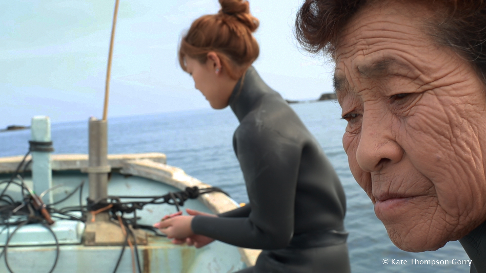 Two women in wetsuits on a boat, with water in the background. The older woman in the foreground has short hair and wrinkled skin, looking contemplative. The younger woman in the background has tied hair and is handling a tool or object. © Kate Thompson-Gorry.
