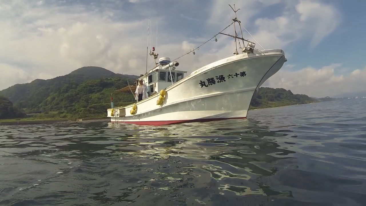 A white fishing boat with Japanese characters on its side floats on calm water near a lush, green hillside under a partly cloudy sky. The boat features antennas and lifebuoys, and a person is visible standing on its deck.