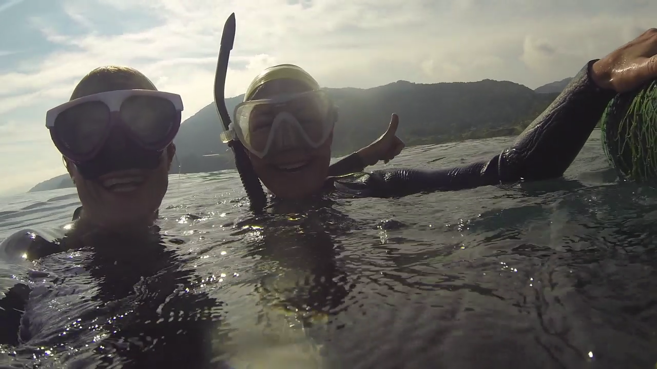 Two snorkelers in wetsuits with masks and snorkels float in the water, smiling. One person gives a thumbs-up. They are in an expansive body of water with mountainous terrain visible in the background under a cloudy sky.