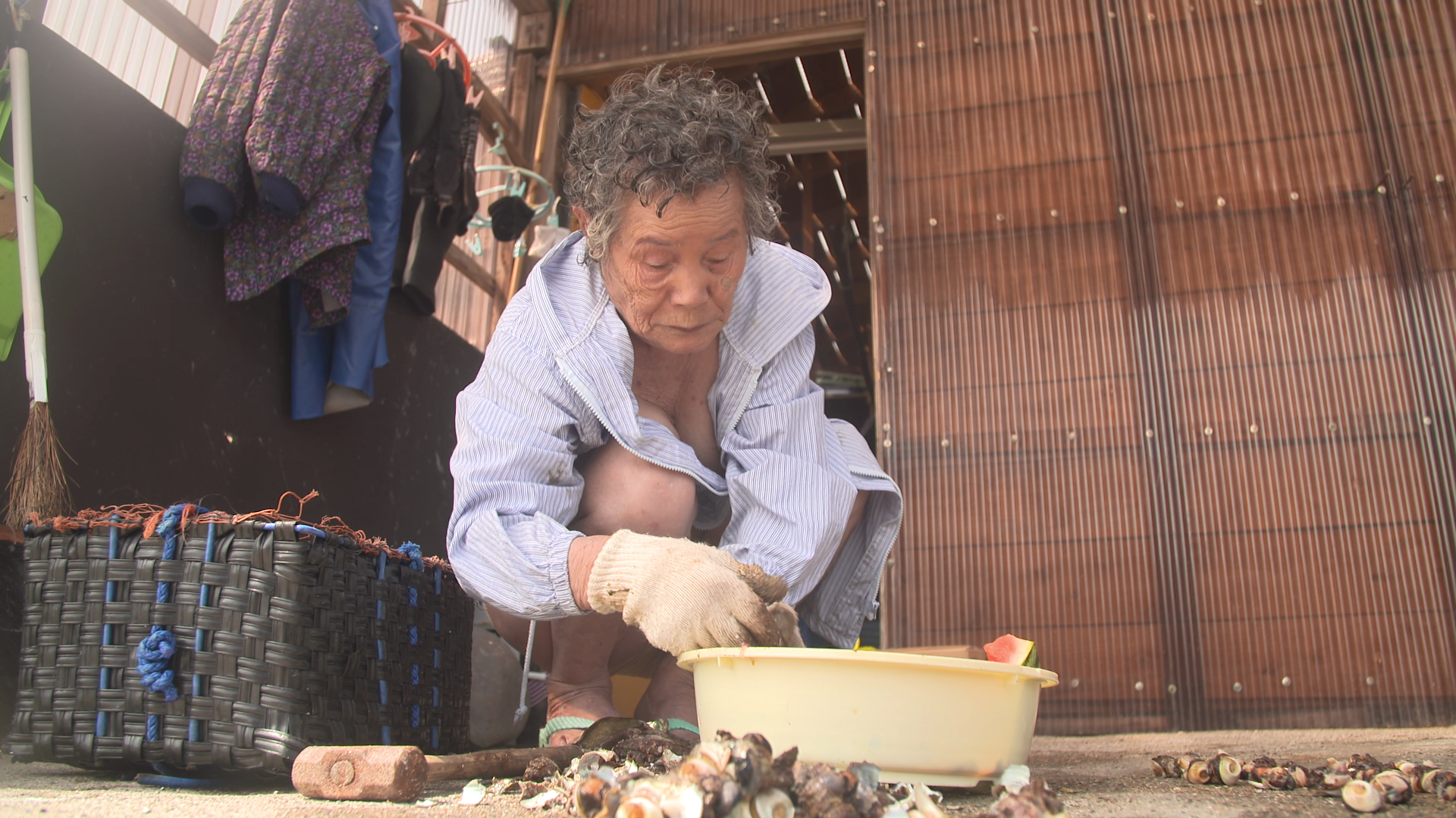 An elderly person crouches in front of a wooden structure, wearing a striped shirt and gloves, sorting sea creatures in a yellow basin. A woven basket and various tools are nearby. Clothing hangs in the background.