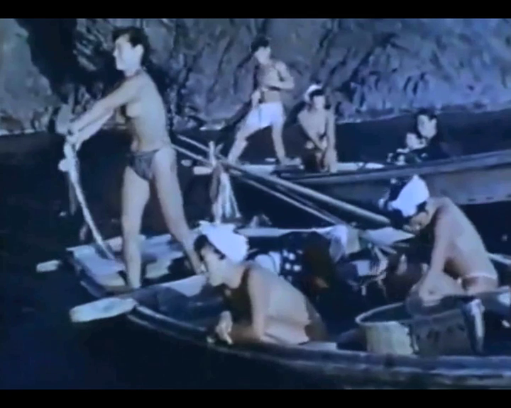 Several people in traditional attire are on small wooden boats, paddling and tending to their tasks. They appear to be in a rocky, coastal area. Some are standing and rowing, while others are seated, focusing on the content in or near the boats.