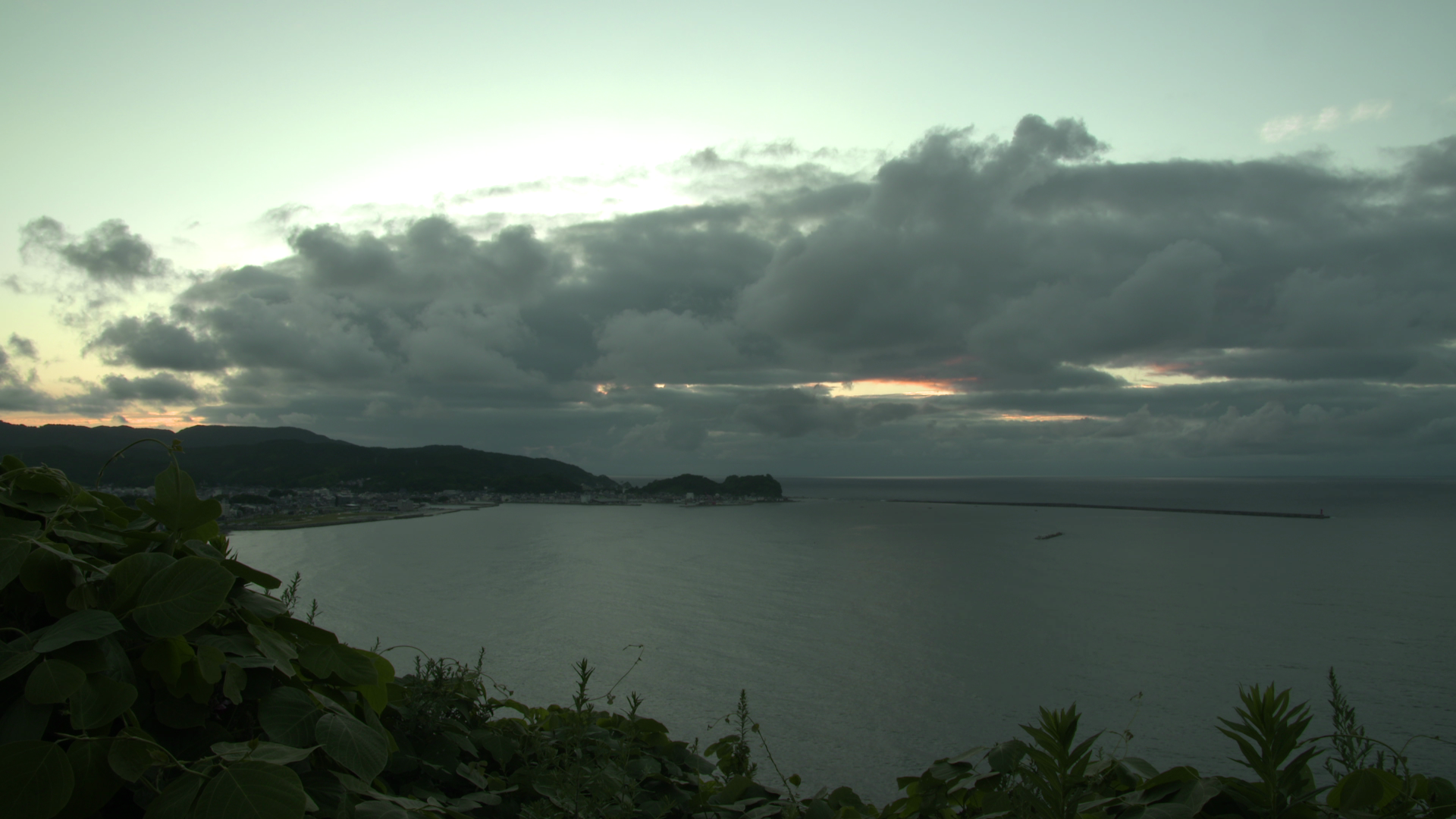 View of a serene coastal landscape at dusk. Large, dark clouds hang in the sky, partially obscuring the fading sunset. Calm waters stretch out to the horizon, bordered by a silhouette of hilly terrain and lush green vegetation in the foreground.