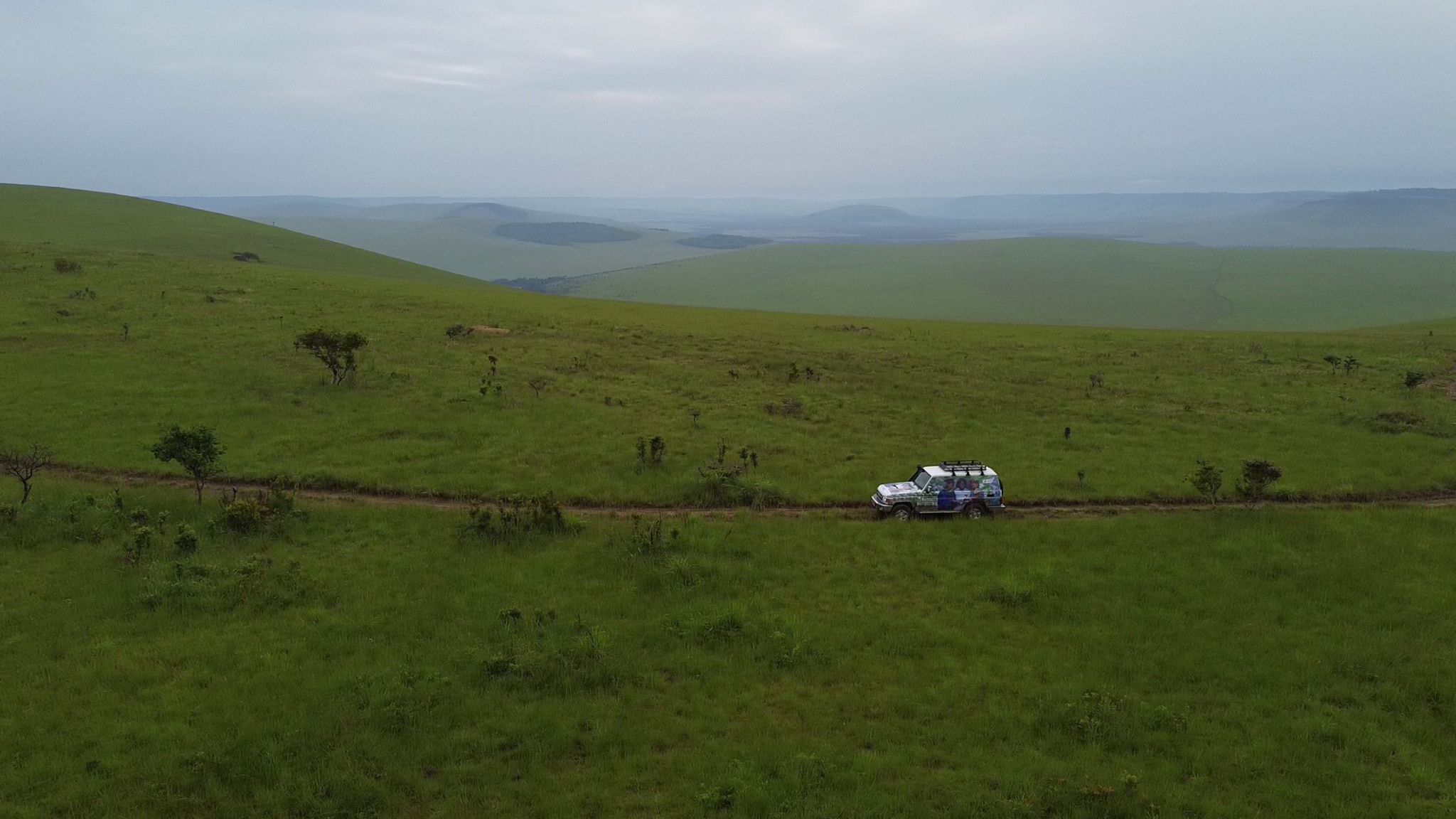 A solitary vehicle with roof cargo drives along a narrow dirt path through vast, rolling green hills under a cloudy sky. Distant hills and valleys stretch out to the horizon, creating a serene, expansive landscape.