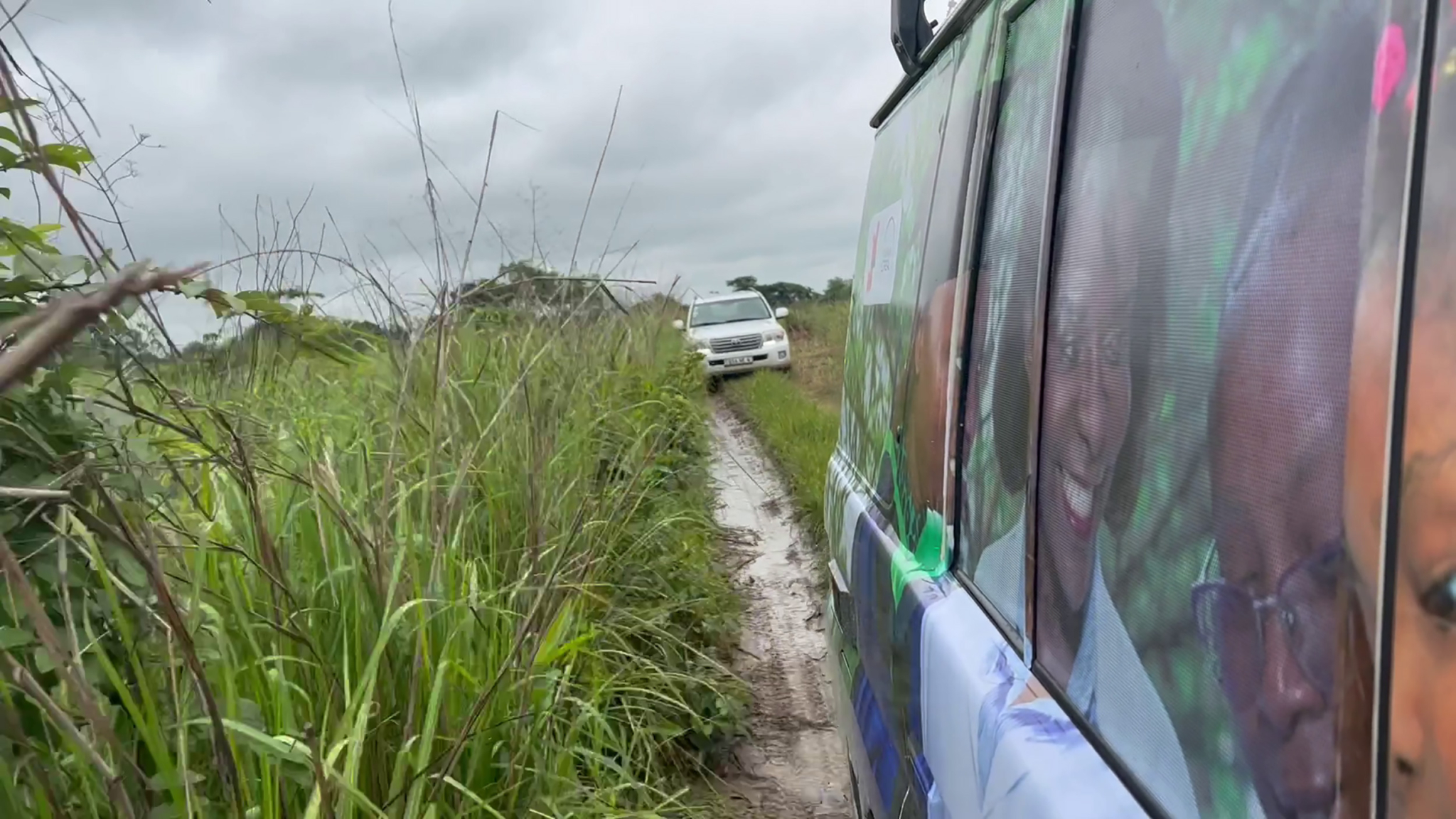 Two vehicles navigate a narrow, muddy, and grassy road under a cloudy sky. The foreground shows the side of a vehicle with a colorful design, while in the background, a white SUV follows closely. Tall grass surrounds the road, suggesting a remote or rural location.