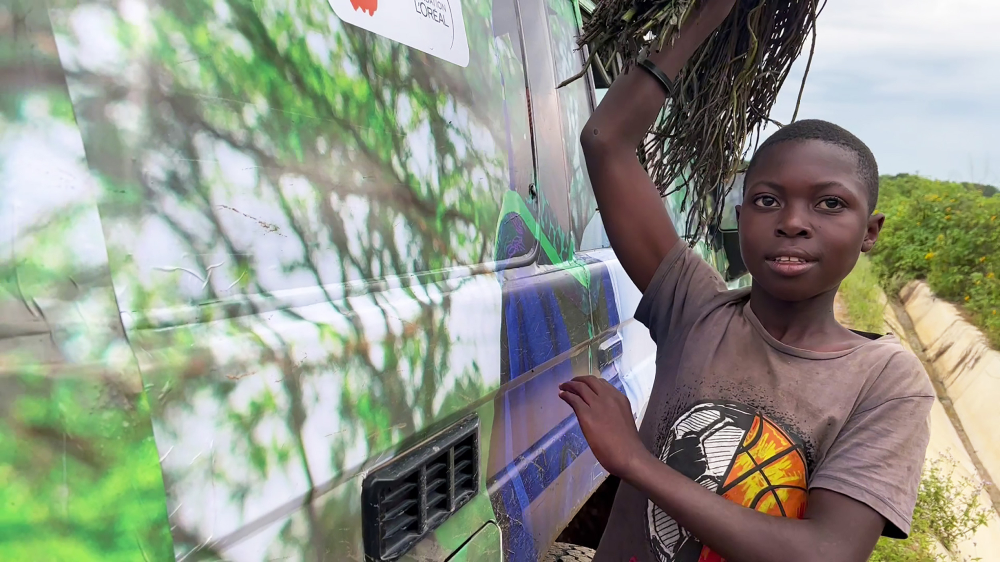 A young person with short hair carries a bundle of sticks on their head while standing next to a colorful vehicle painted with green, white, and blue patterns. They wear a graphic t-shirt and have an arm extended towards the vehicle. Trees are visible in the background.