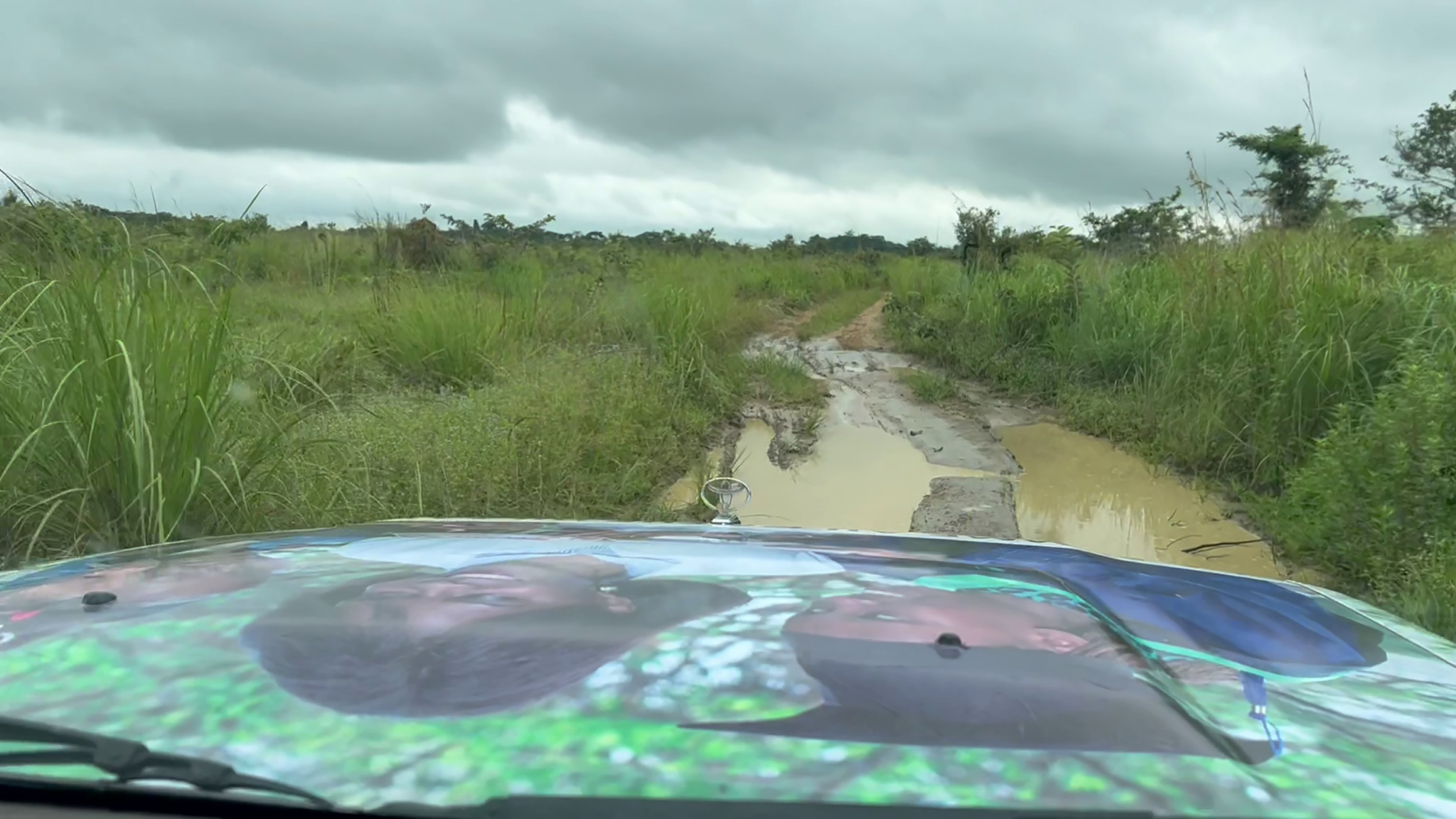 View from the front of a vehicle traveling down a muddy, rural dirt road surrounded by green grass and bushes. The vehicle's hood features a large image of two people, partially visible. The sky is cloudy, hinting at possible rain.