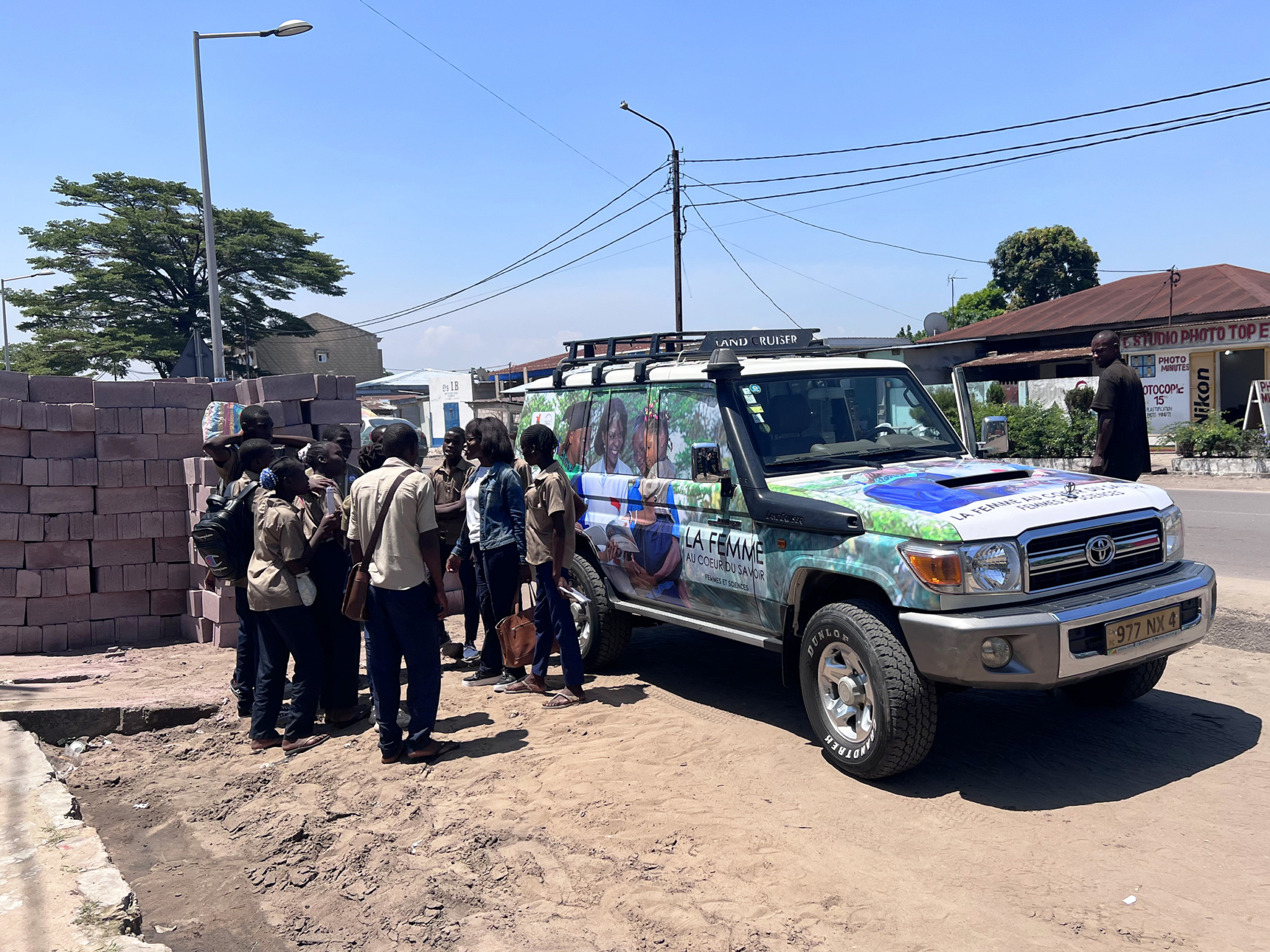A group of people, including several individuals wearing tan shirts, gather around a colorful SUV parked by the roadside on a sunny day. The SUV has vibrant graphics on it and a roof rack. In the background are stacked bricks, street signage, and utility poles.