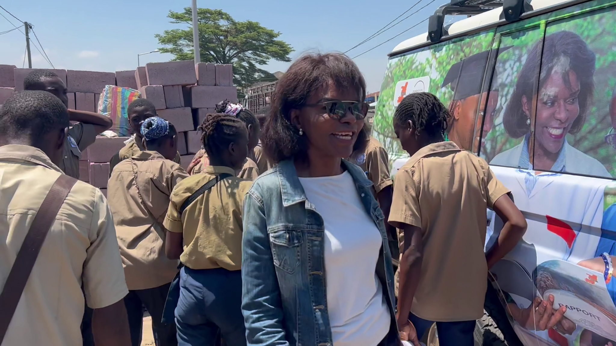 A woman in a denim jacket and sunglasses smiles as she walks among a group of schoolchildren dressed in uniforms. The children surround a vehicle decorated with colorful designs and images. There are stacks of bricks in the background. The scene is outdoors on a sunny day.