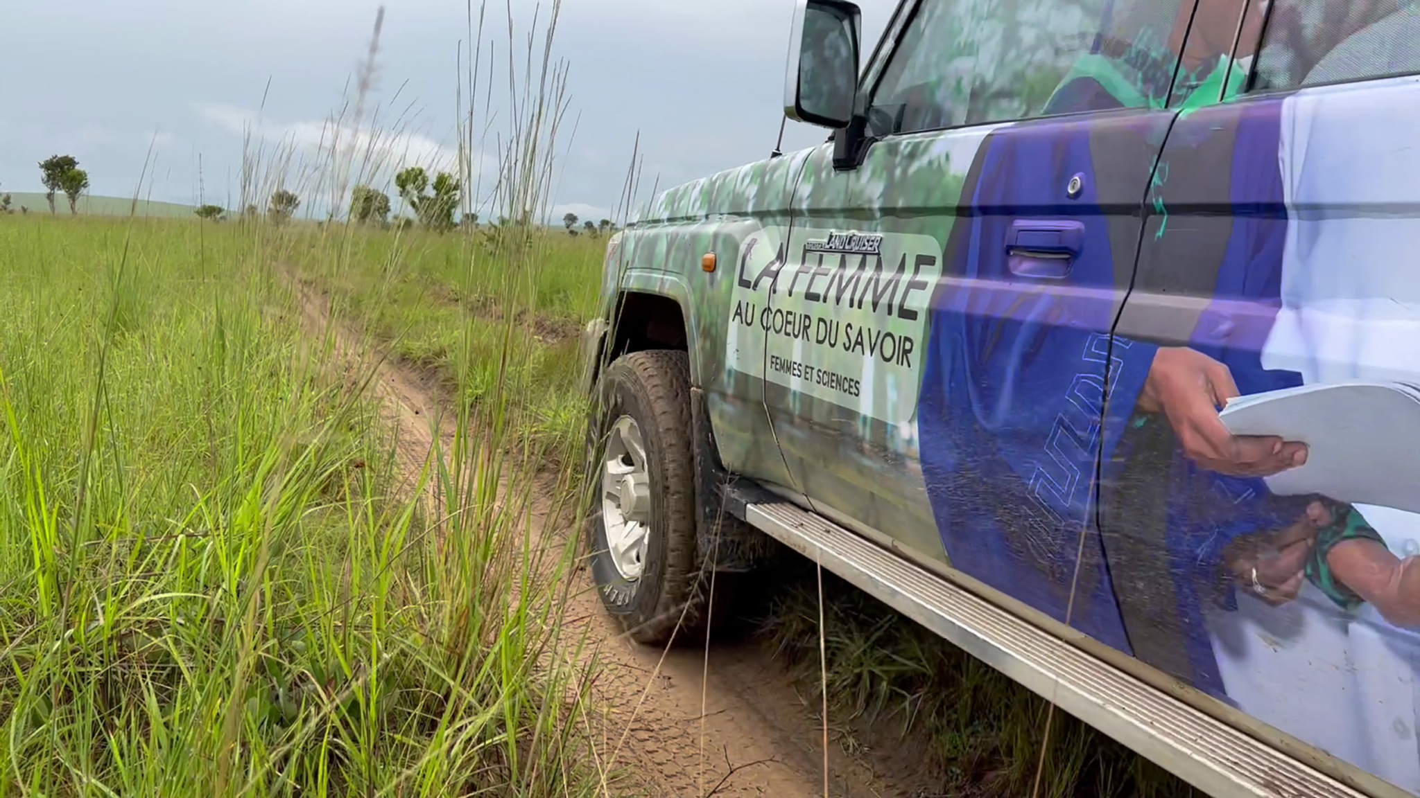 A 4x4 vehicle drives on a narrow dirt path through a grassy field under a cloudy sky. The vehicle's side bears a large decal with French text partially visible, showing "LA FEMME AU COEUR DU SAVOIR." Tall grass surrounds the route and stretches into the distance.