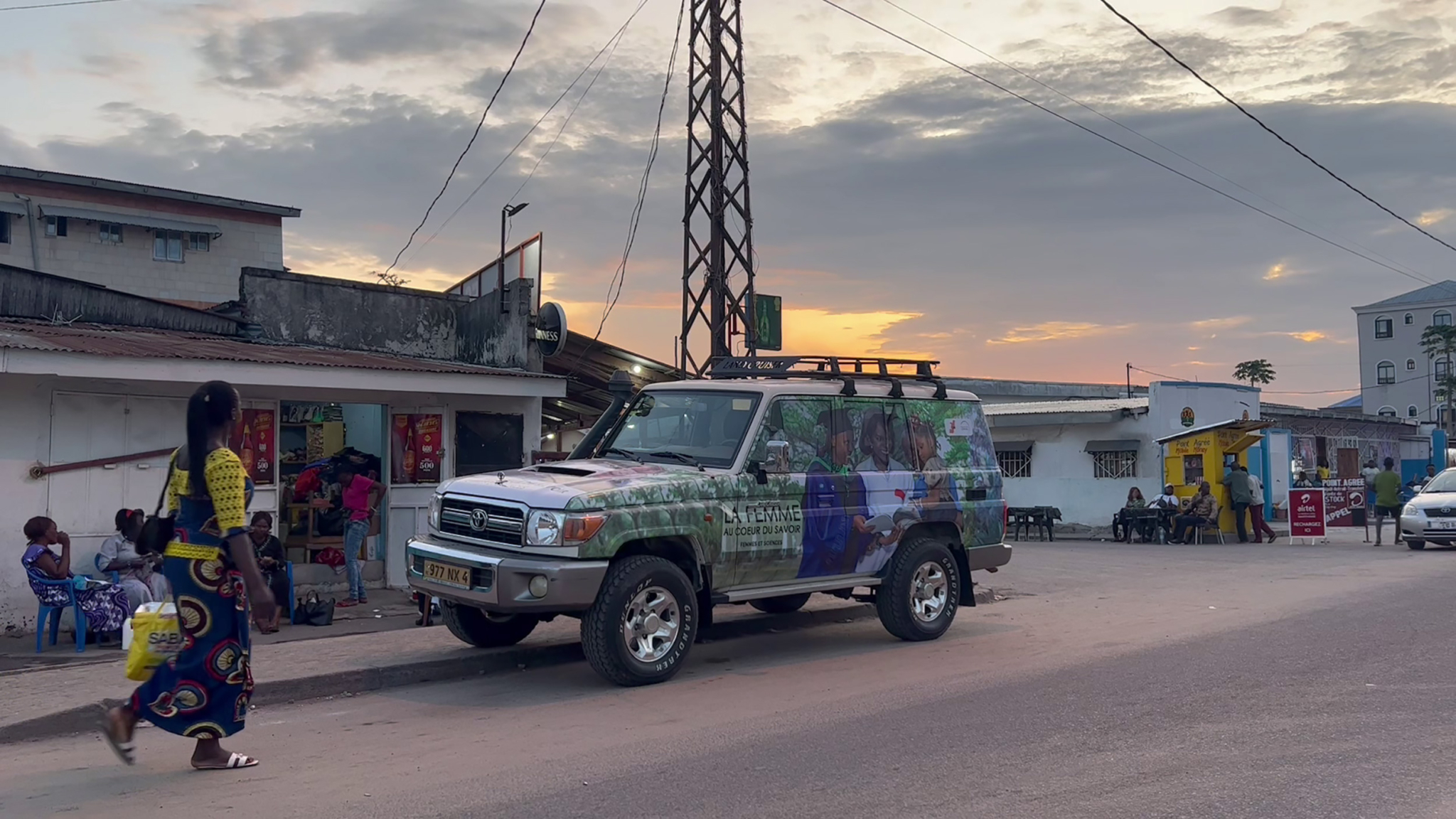 A street at sunset with a painted SUV parked on the side. People are seen walking and sitting outside nearby buildings. Power lines and a pole tower in the background, with a partly cloudy sky adding a warm glow to the scene.