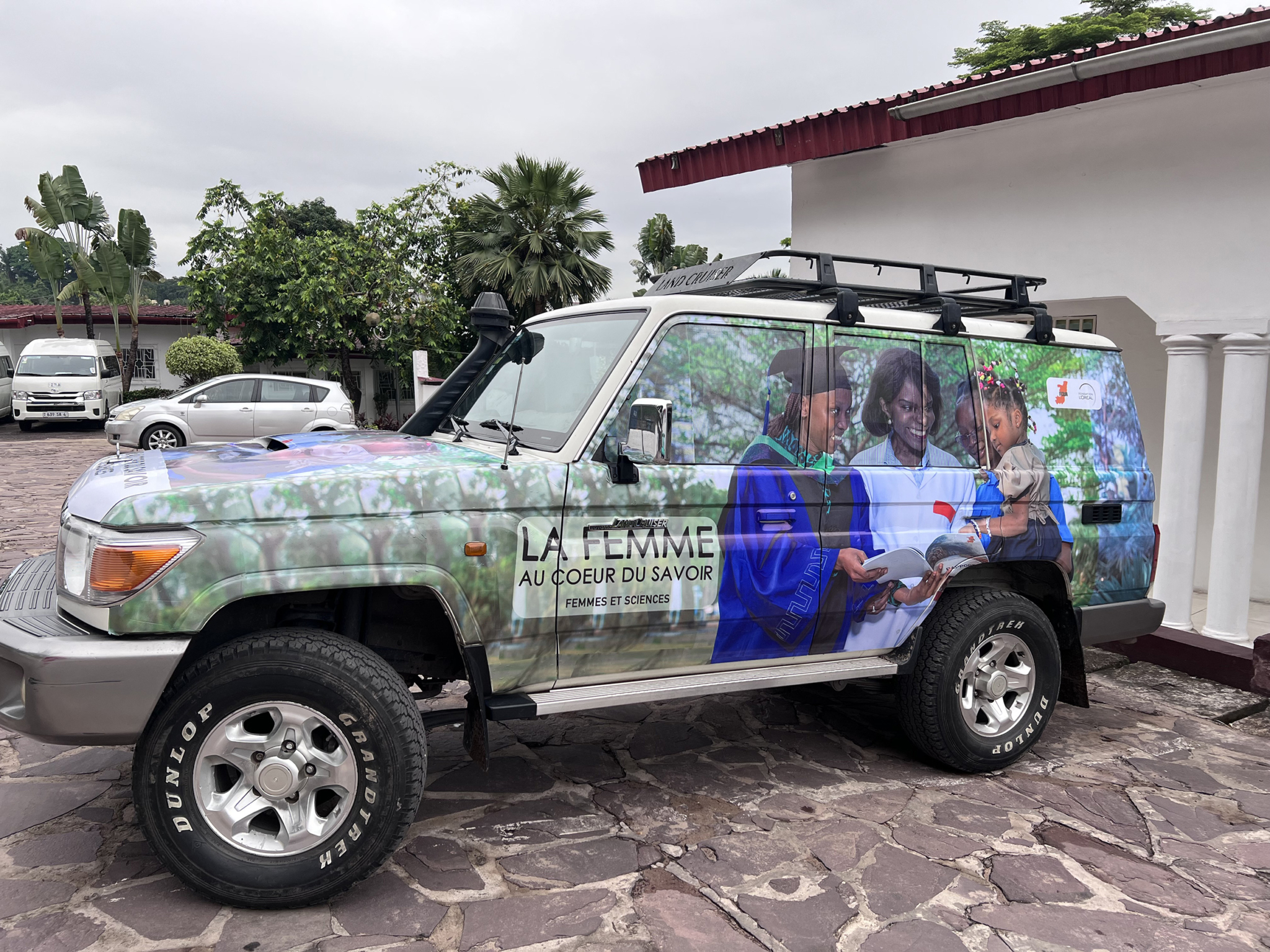 A white SUV displays a large wrap featuring an image of three women: one in a graduation gown, one in a lab coat, and a young girl holding a book. The text reads "LA FEMME AU COEUR DU SAVOIR" and "FEMMES ET SCIENCES." The vehicle is parked on a stone pavement.