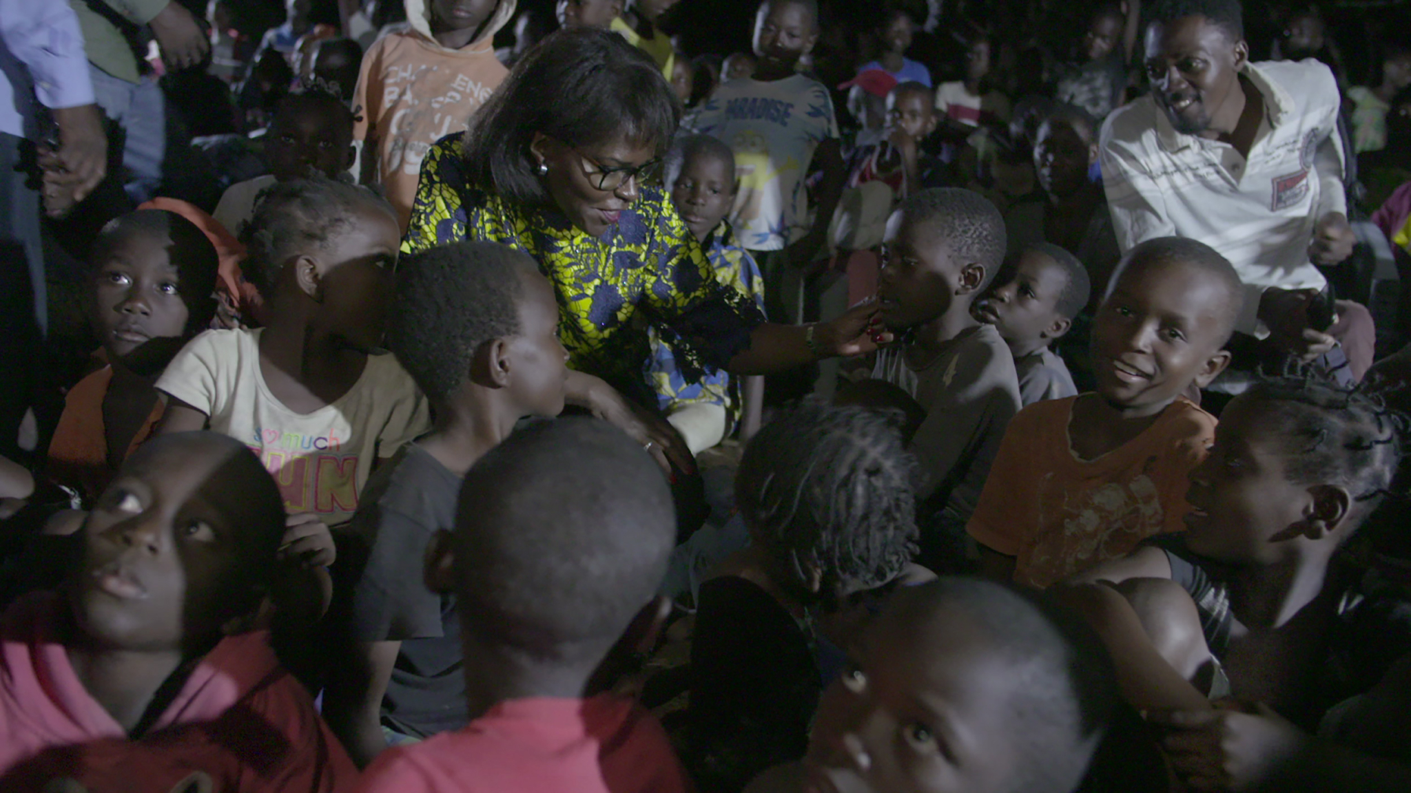 A woman wearing glasses and a colorful shirt is surrounded by a group of children in a dimly lit environment. She is engaging with the children, smiling and placing her hand gently on one child's face. All the children are looking at her, some smiling.