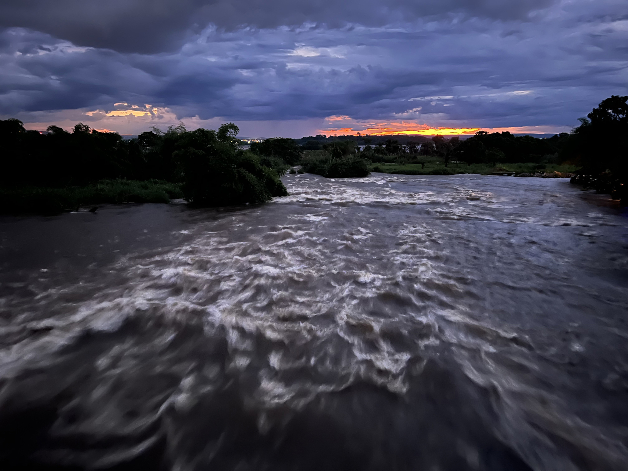 A turbulent river flows under a dramatic evening sky with thick clouds. The horizon reveals a vibrant, glowing sunset in the distance, contrasting the dark, cloudy sky above. Trees and foliage line the riverbanks, silhouetted against the colorful sunset.
