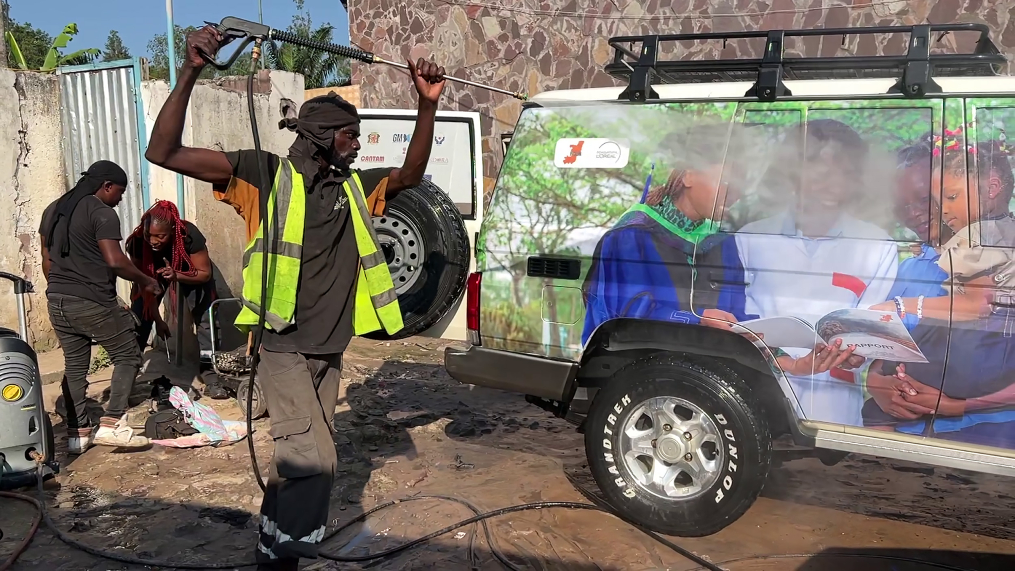 Two people cleaning the outside of an off-road vehicle with a hose and scrubbing brush. The vehicle is muddy, and one person in a reflective safety vest and cap raises their hands while spraying water. The car features an image of children on the side.