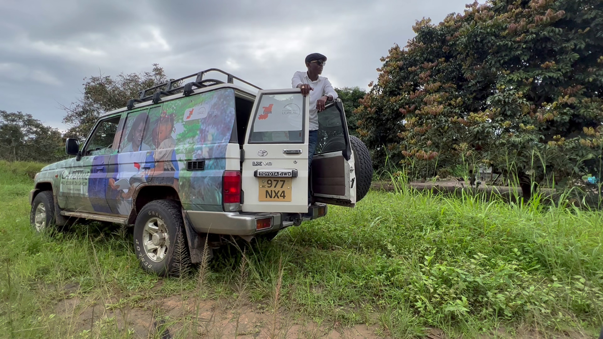 A person stands at the back of a Land Cruiser SUV, which has a colorful exterior adorned with images and a roof rack. The vehicle is parked on a grassy area with lush green trees in the background under a cloudy sky. The license plate reads "977 NX4.