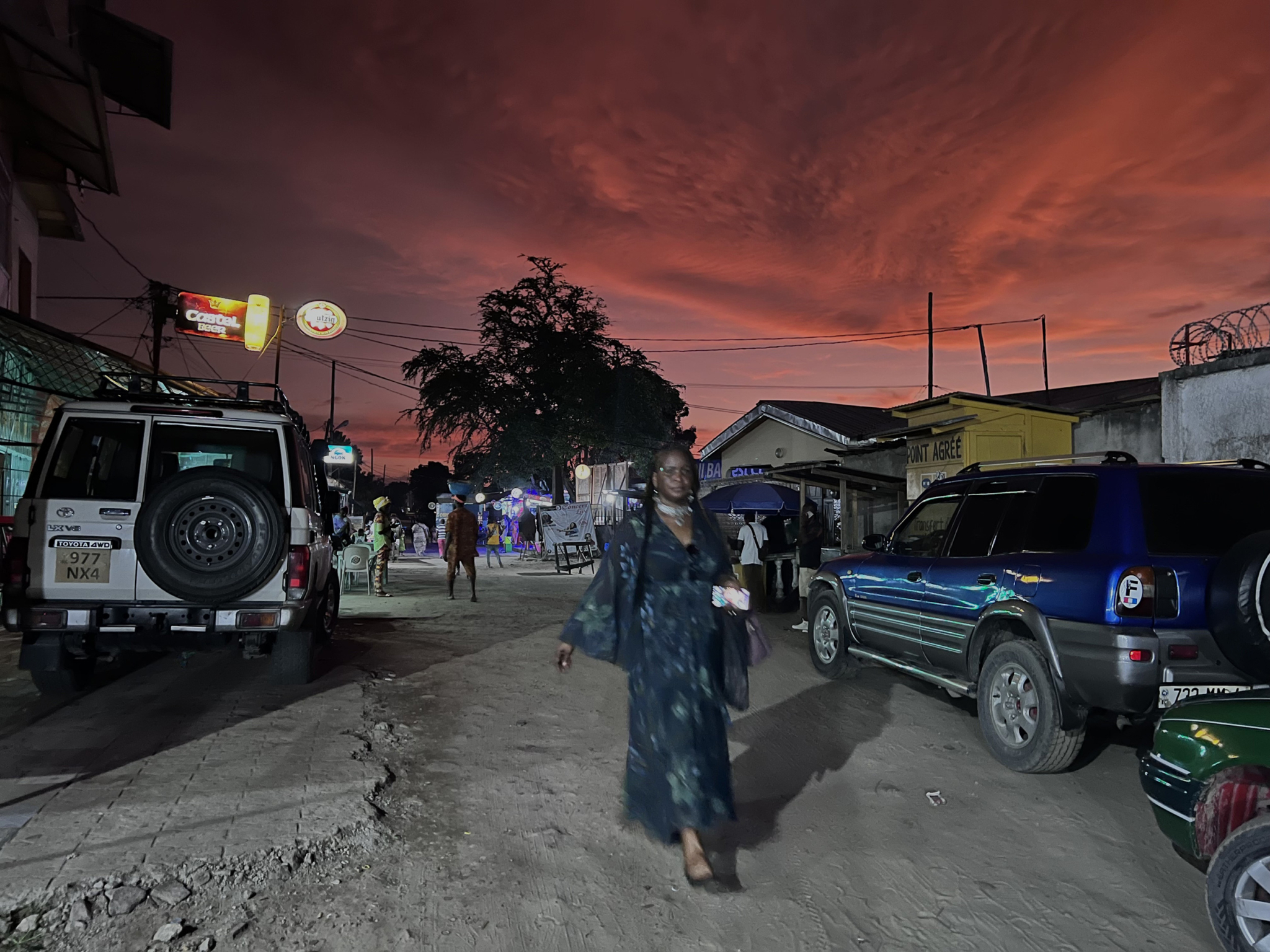A woman wearing a long dark dress walks down a street at dusk. The sky is a mix of dark and bright red hues. Cars are parked along the sides, and people are visible in the background near lit storefronts. The atmosphere has a busy, yet calm, evening vibe.
