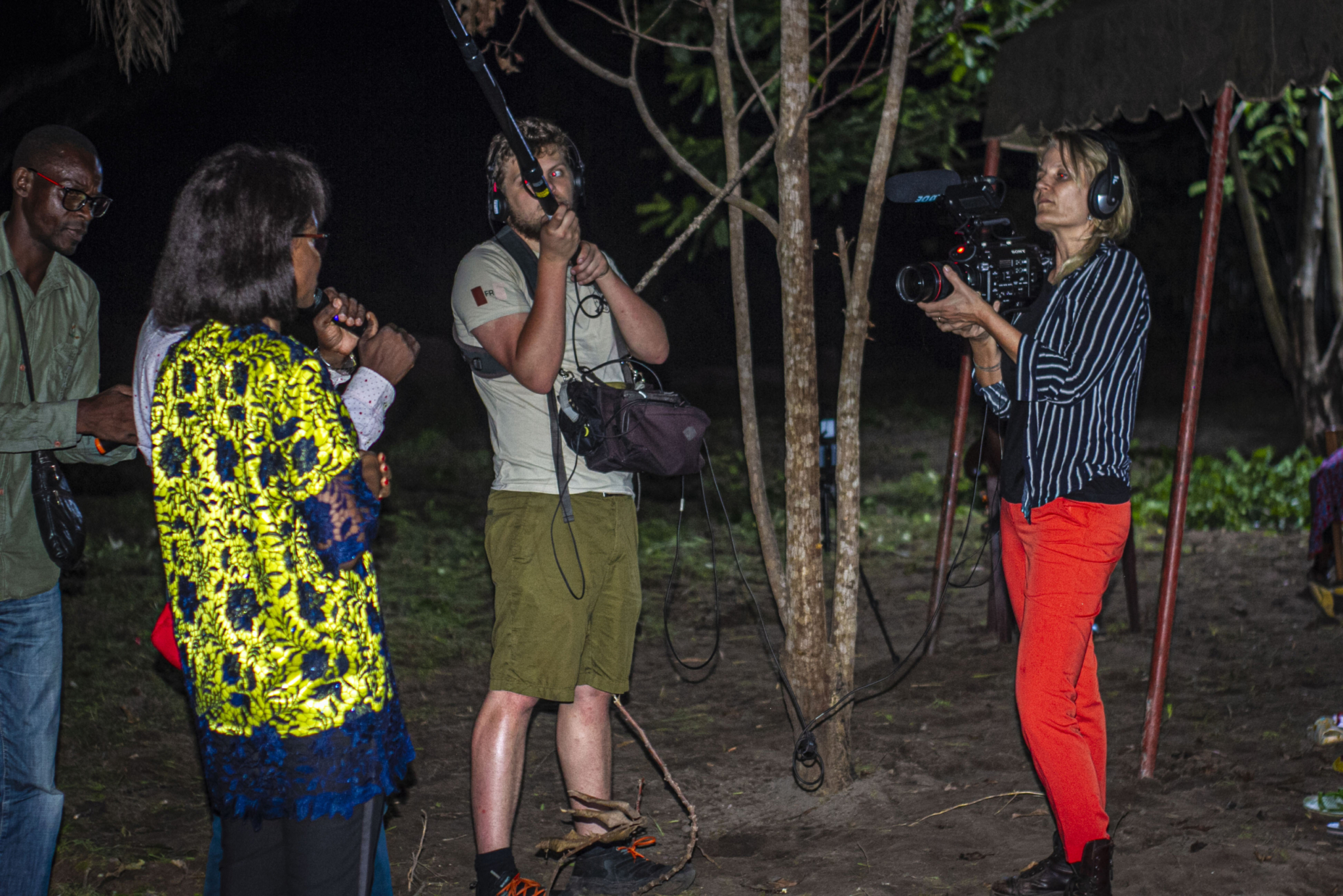 A woman wearing a striped shirt and red pants films with a camera as two individuals hold boom microphones. Another person in a vibrant yellow and blue shirt stands speaking to the camera. The scene appears to be taking place outdoors at night.