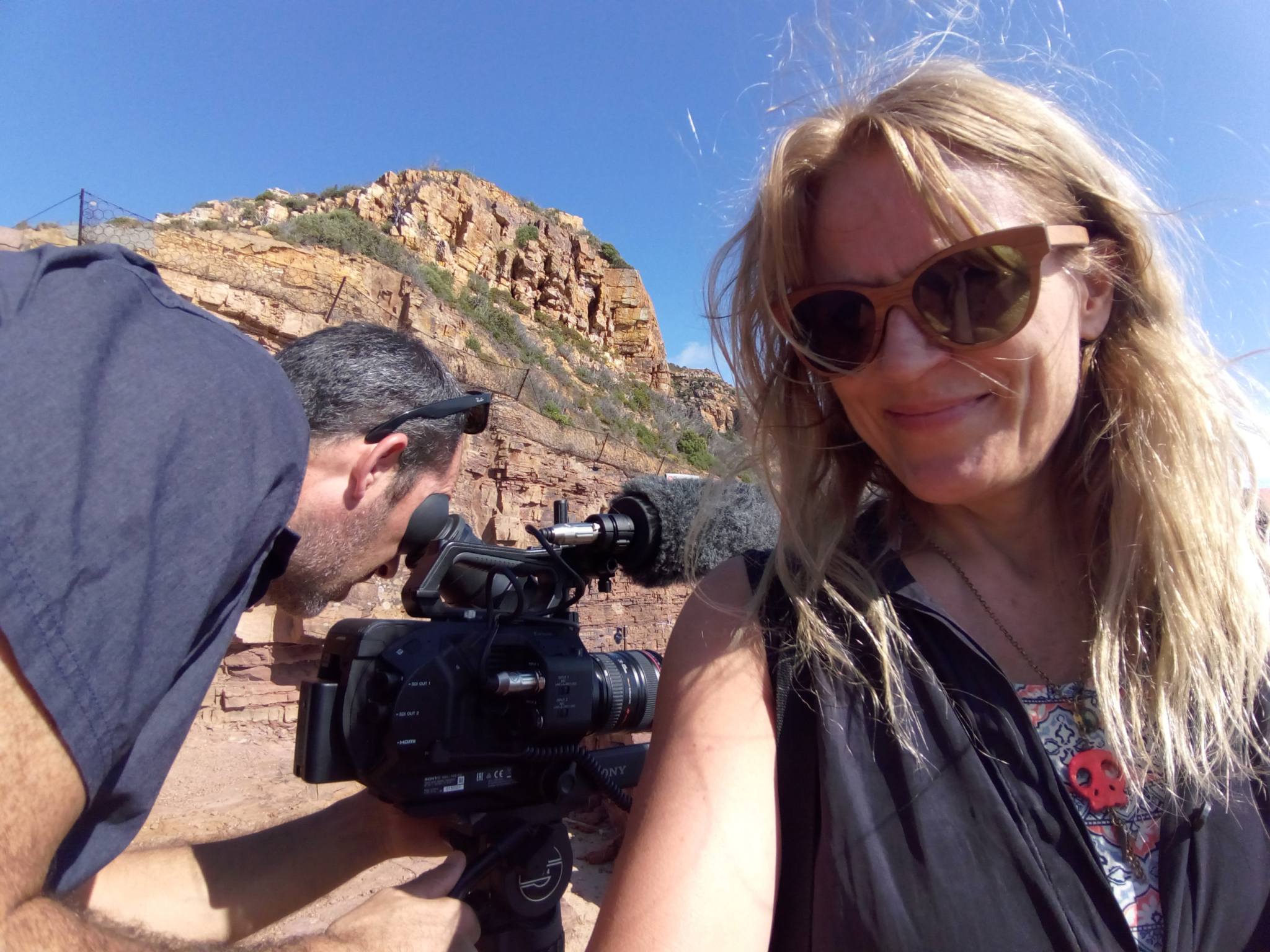 A woman wearing sunglasses smiles at the camera, while a man operates a video camera beside her. They are outdoors near a rocky landscape with clear blue skies.