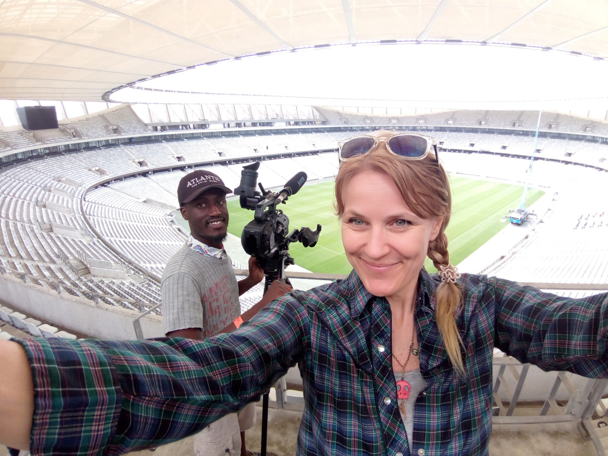 Two people are taking a selfie in a large, empty sports stadium. The woman in the foreground, with sunglasses and a plaid shirt, holds the camera. The man behind her smiles and stands next to a professional video camera on a tripod. The field is visible in the background.