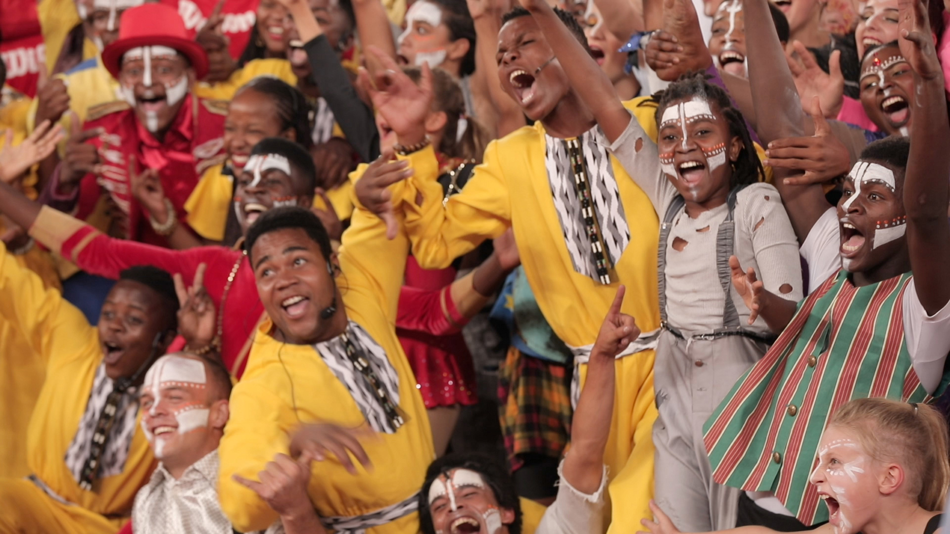 A lively group of people adorned in colorful outfits, face paint, and various costumes joyfully celebrates. They are energetically waving, cheering, and smiling, creating an atmosphere of excitement and unity.