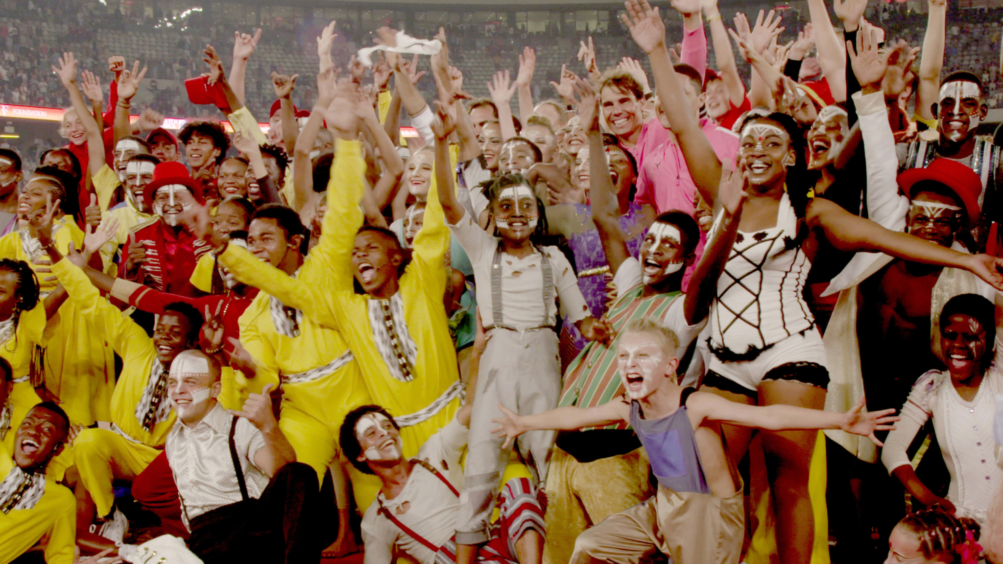 A large, diverse group of performers dressed in colorful and elaborate circus costumes celebrate together in a stadium. They are variously painted with face paint and wear intricate outfits. Many have their hands raised in joy, and the crowd behind them is blurred.