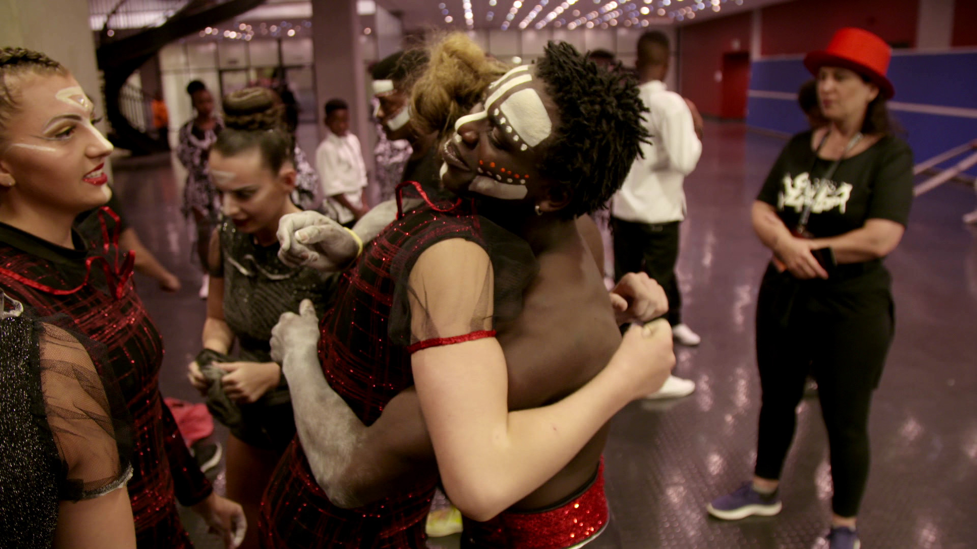 A group of performers is backstage in an embrace, with two people in the foreground sharing a hug. They have theatrical makeup and vibrant costumes. In the background, other performers and individuals are engaging in conversation. The setting appears to be a hallway.