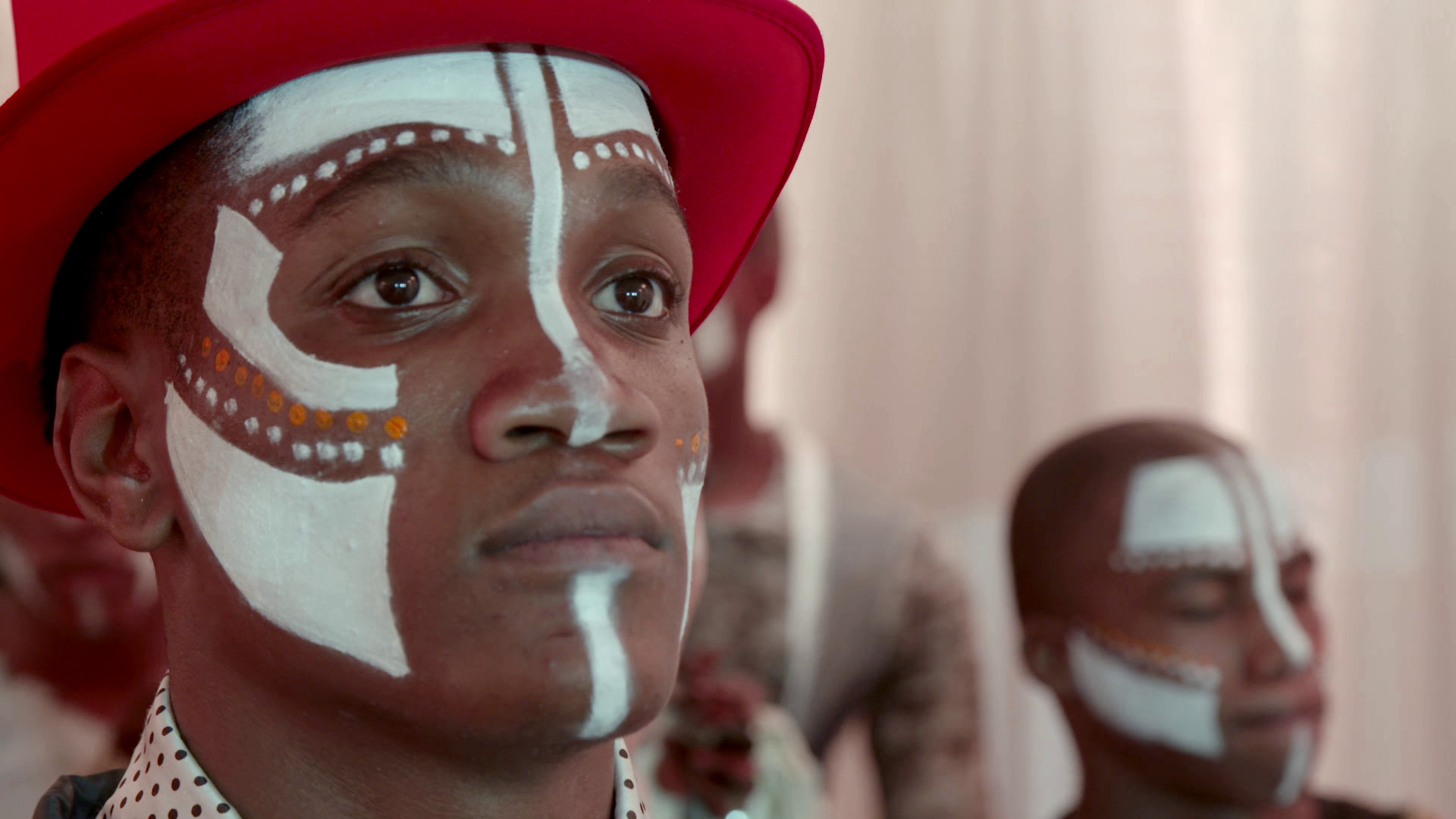 A person wearing a bright red hat with traditional white face paint and small orange dots looks forward with a serious expression. In the background, another person with similar face paint is partially visible.