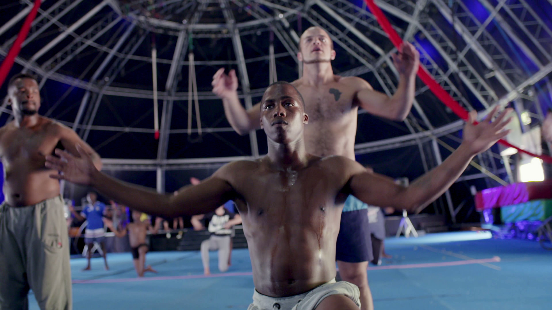 A group of shirtless men practice acrobatics in a large indoor facility with a dome-like structure overhead. The man in the foreground kneels with arms extended, while others perform synchronized movements behind him. The scene suggests a circus or gymnastic training.