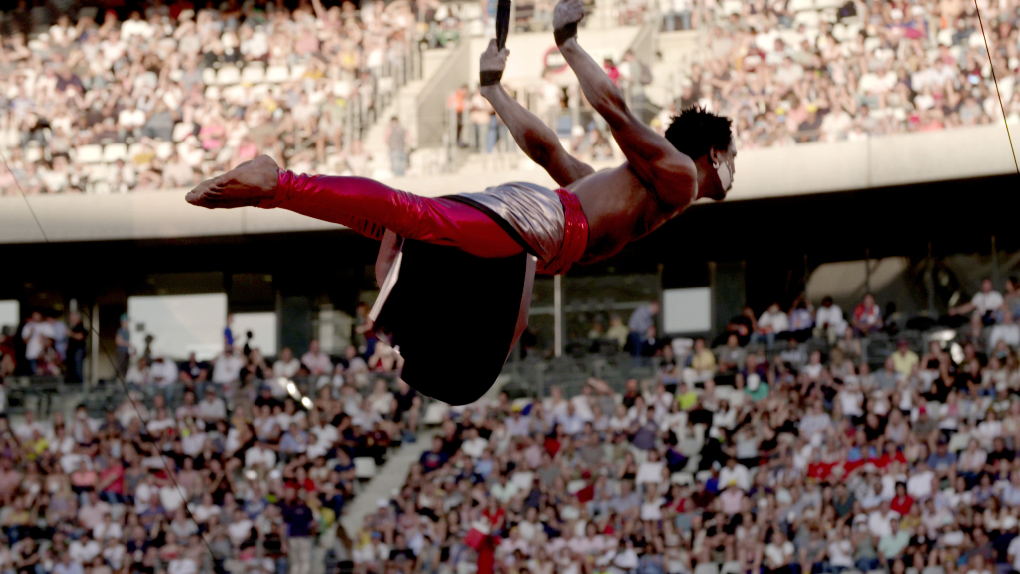 An acrobat in red pants and a shiny silver belt performs on aerial rings, suspended high above a crowded stadium audience during daylight.