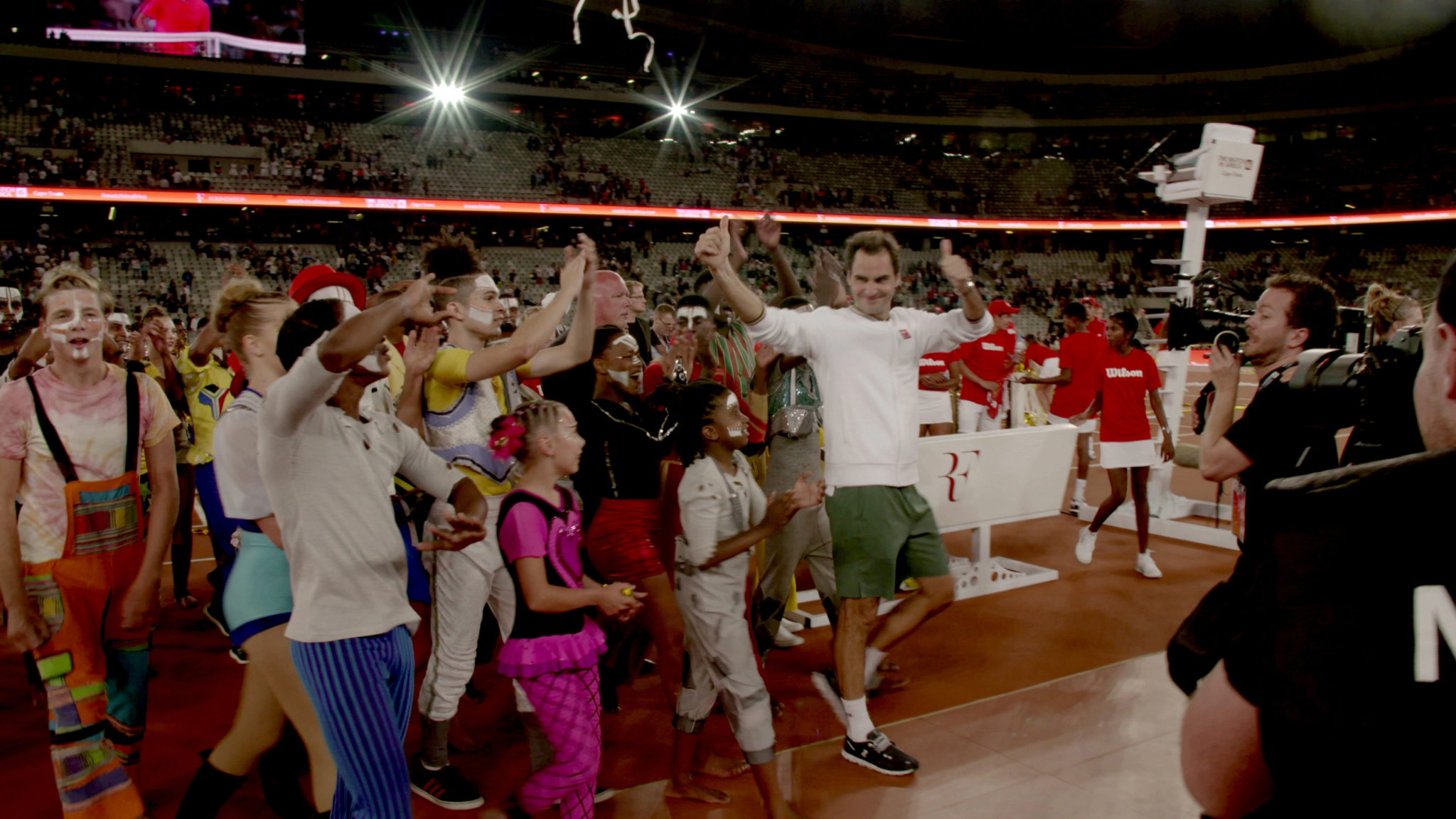 A crowd of people joyfully celebrating on a sports field. The group includes both adults and children, some dressed in colorful costumes and clown outfits. They are gathered around a man in a white shirt and green shorts, who appears to be the focus of the celebration.