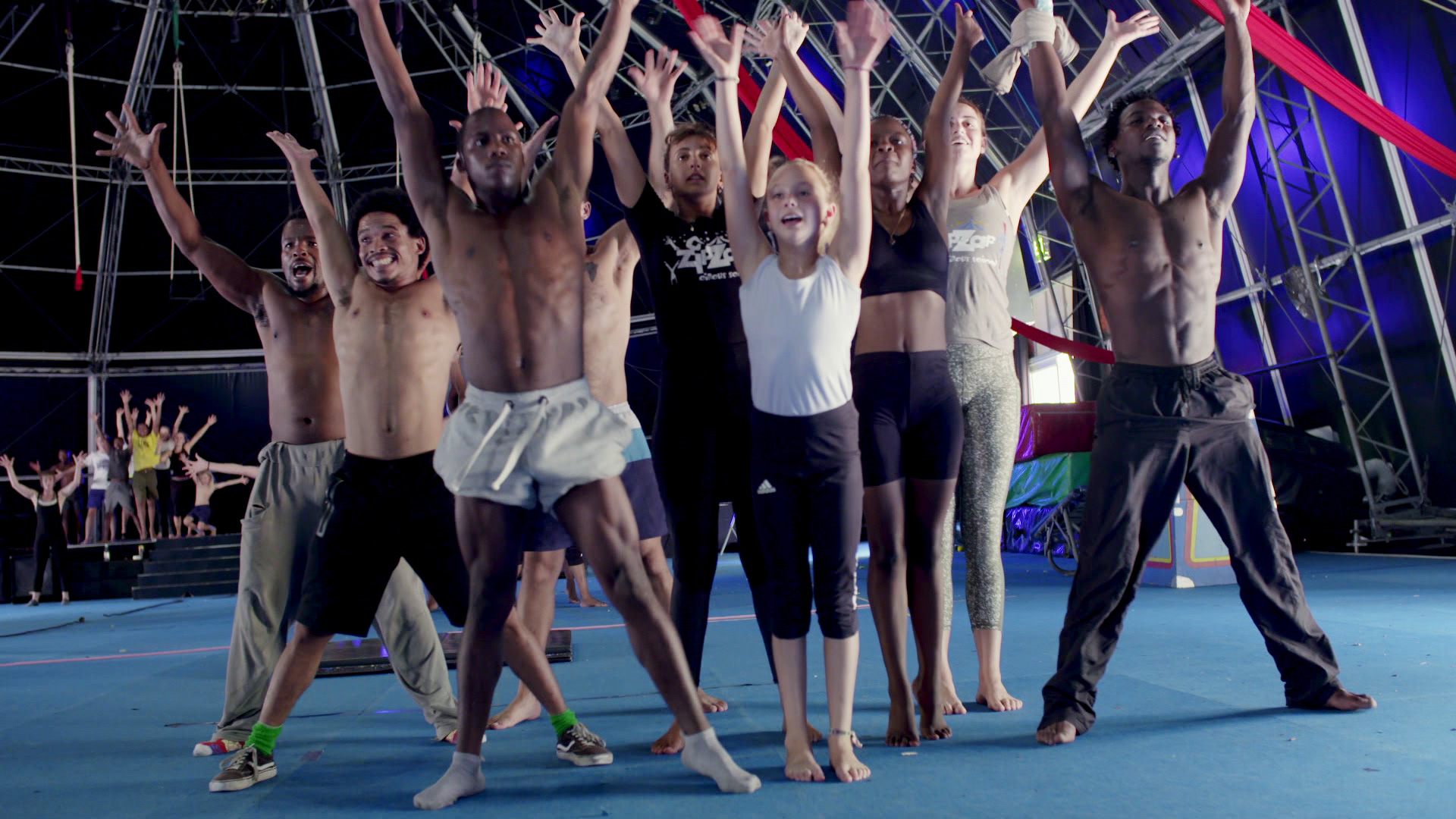 A group of people dressed in athletic clothing are enthusiastically raising their arms while standing on a blue floor in what appears to be a circus or performance space, with a high ceiling structure and various equipment visible in the background.