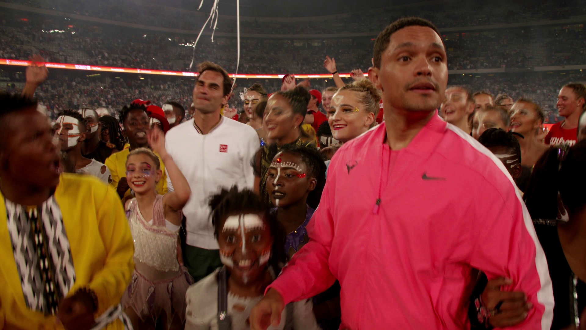 A group of people, including children with face paint, celebrate together. A man in a pink jacket stands in the foreground, while another man in a white shirt is behind him. They're surrounded by a joyous crowd in a stadium.