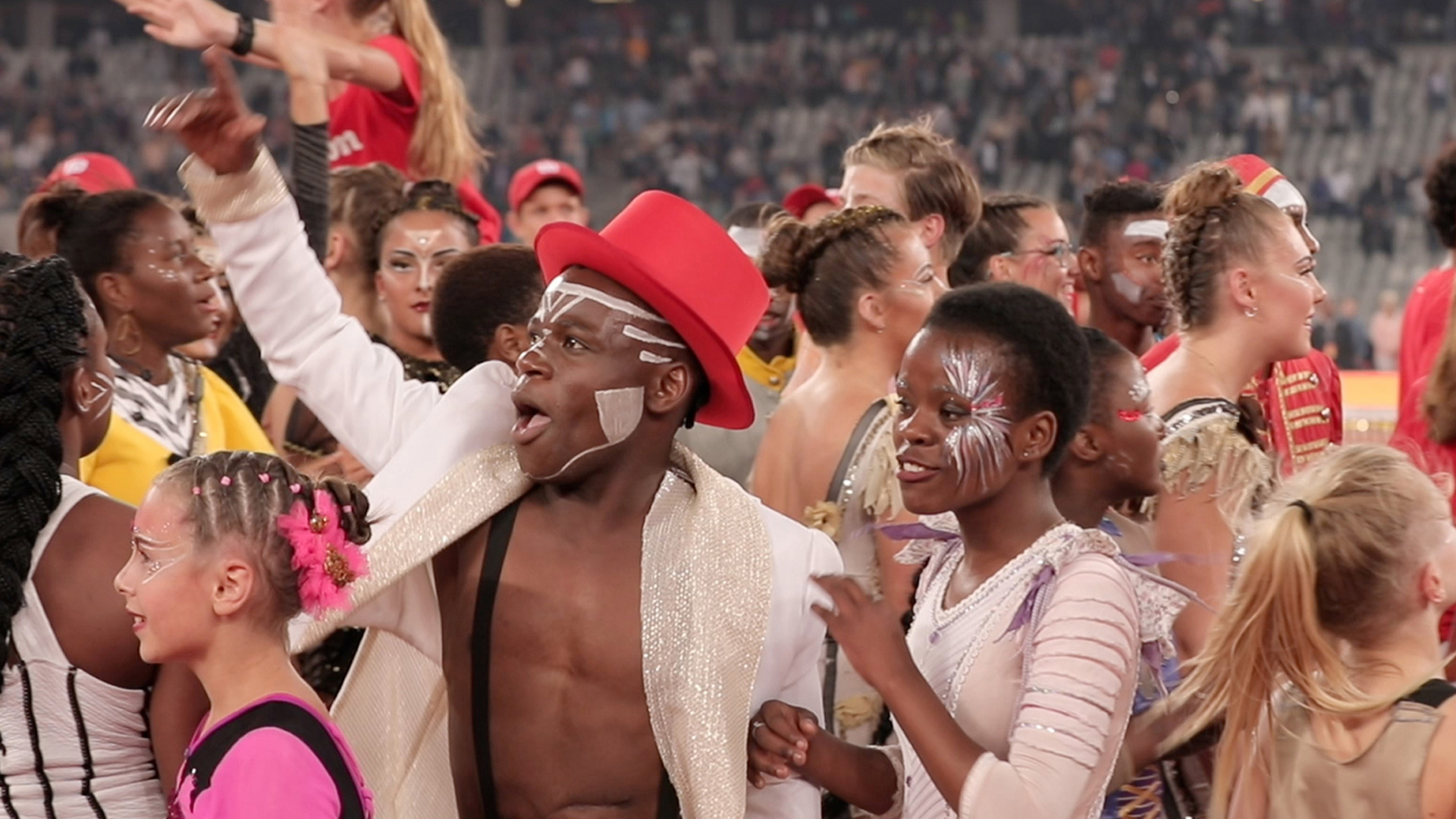 A crowd of people in festive, colorful attire and face paint are celebrating. A man in the foreground, wearing a red hat and a white jacket, points to the distance excitedly, while others around him appear joyful and engaged in the lively atmosphere.