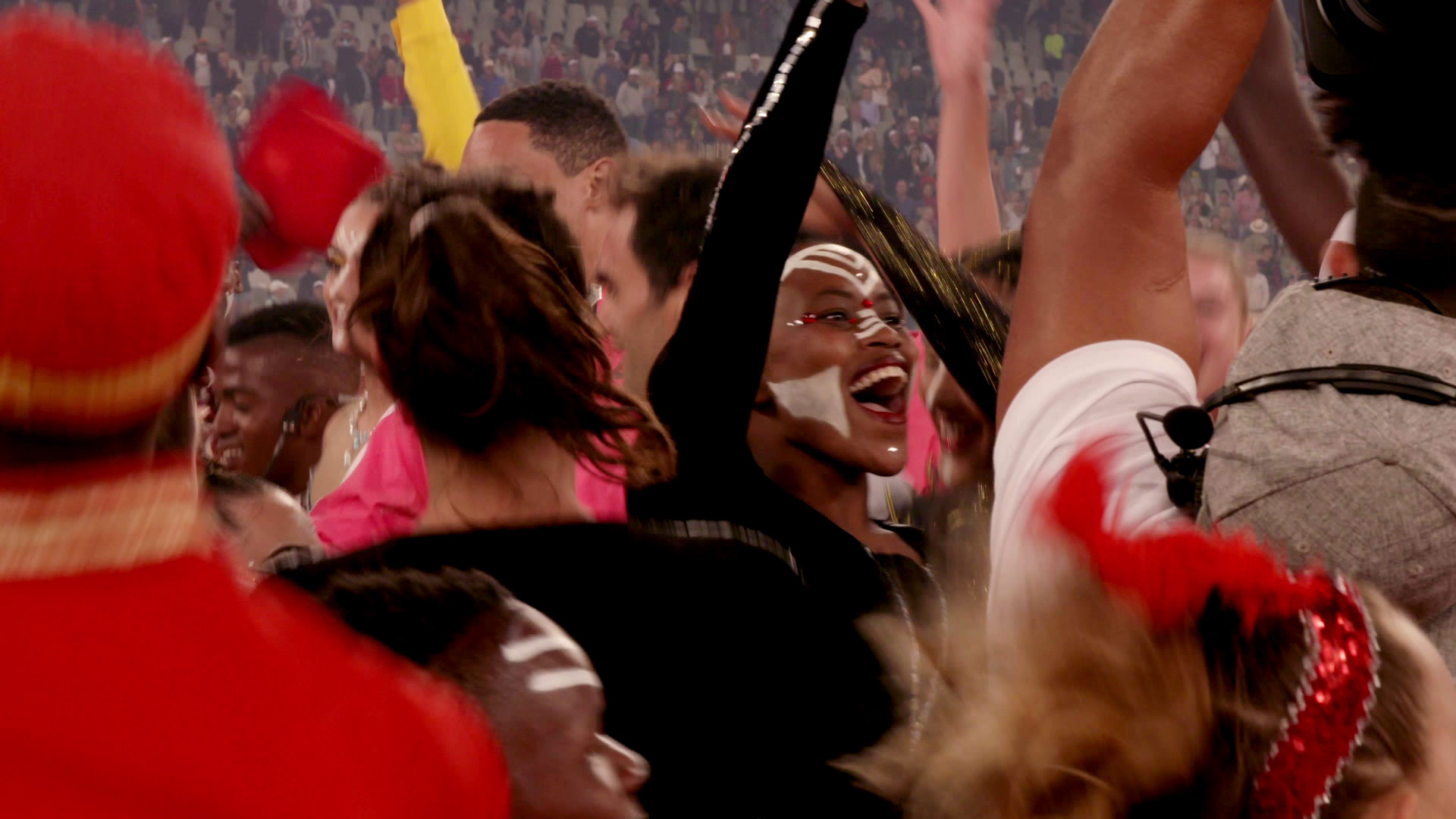 A jubilant crowd is seen celebrating with painted faces and raised arms. The atmosphere is energetic, with many people clustered together, some in vibrant outfits and face paint. The background is filled with more people, indicating a lively, spirited event.