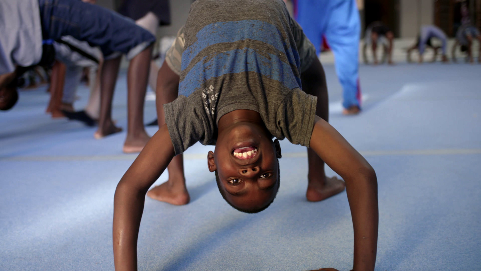 A child doing a backbend during a group exercise session on a blue mat. Others in the background are also participating in various stretching and balancing activities. The child is wearing a grey and blue shirt and smiling upside down at the camera.