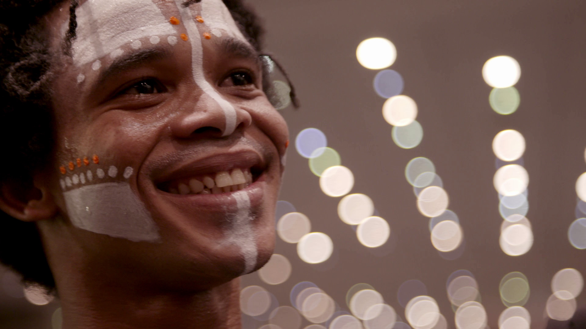 A person with face paint featuring white lines and dots, along with small orange dots, smiles brightly. The background is a blur of circular lights, creating a bokeh effect.