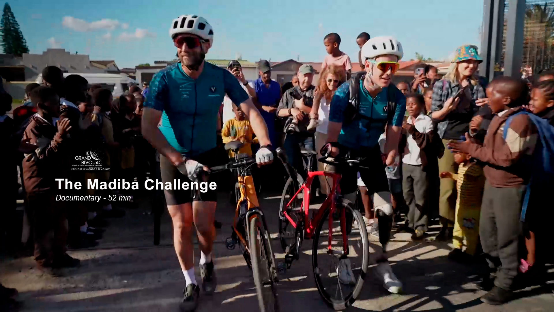 Two cyclists in matching teal jerseys and white helmets walk their bikes through a crowd. Onlookers cheer and take photos. Overlay text reads: “The Madiba Challenge. Documentary - 52 min”. The backdrop includes vibrant community scenes.