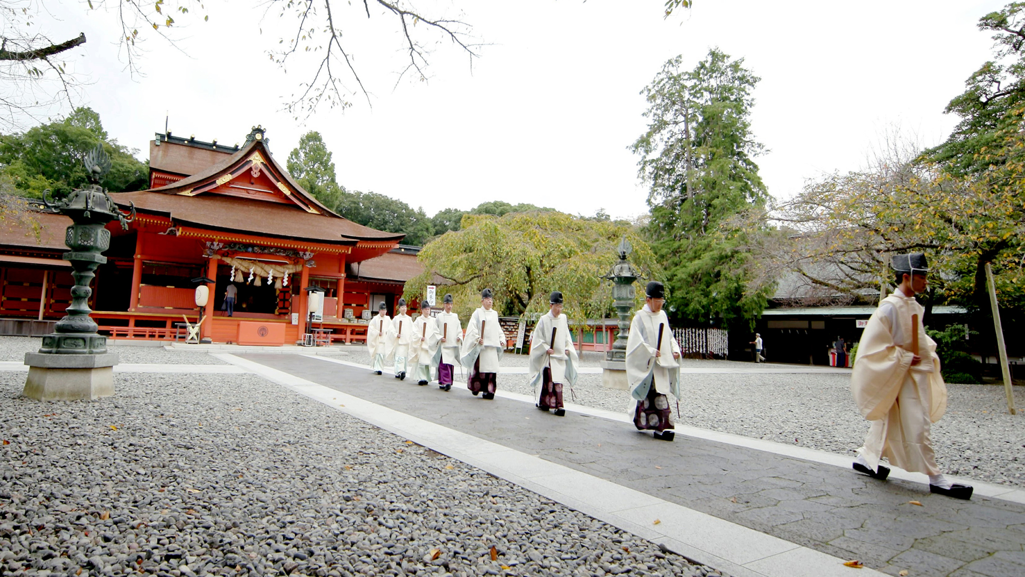 A group of people dressed in traditional Japanese garments walk in a single file towards a historic shrine. The shrine buildings feature red wooden architecture with intricate designs. The surrounding area is serene with stone paths and well-kept trees.