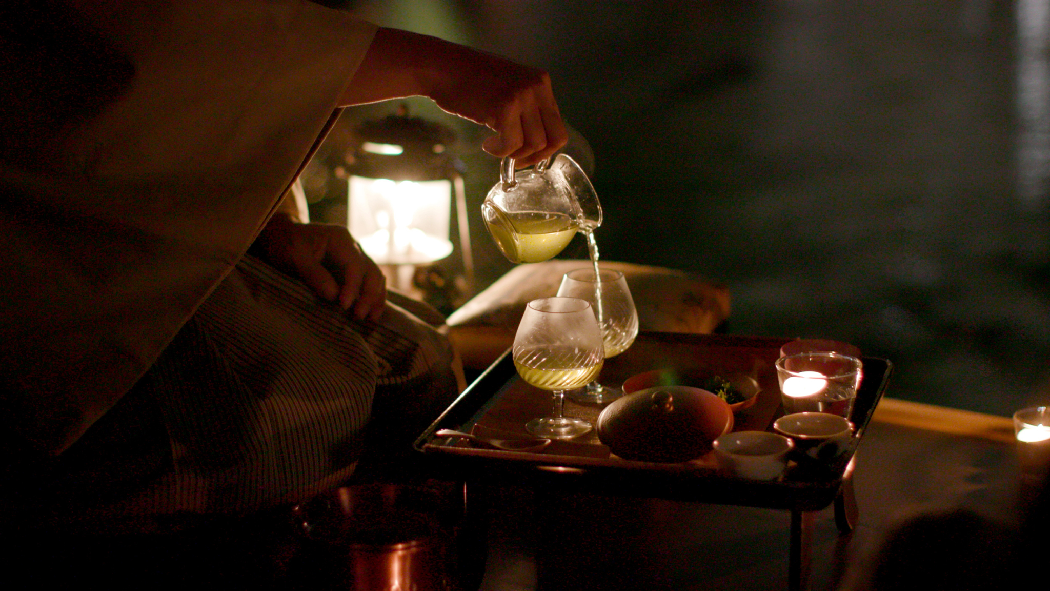 A person pours a light green drink from a small glass pitcher into two glasses on a tray, lit by a soft glow from nearby candles and a lantern. The tray also holds small dishes and fruit, creating a cozy, intimate setting.